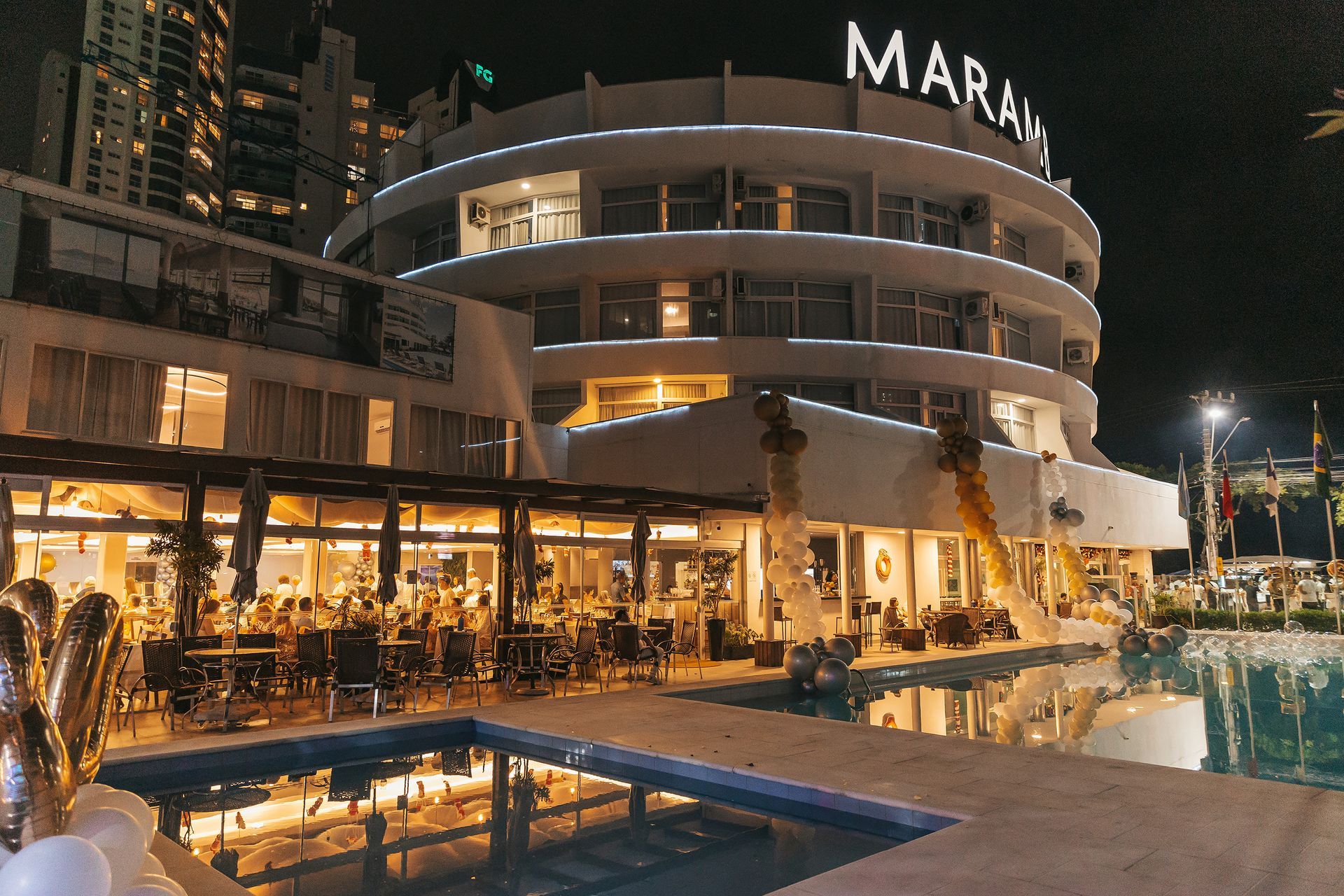 Vista noturna de uma piscina iluminada de um resort e do terraço de um restaurante ao lado de um edifício curvo à beira-mar.