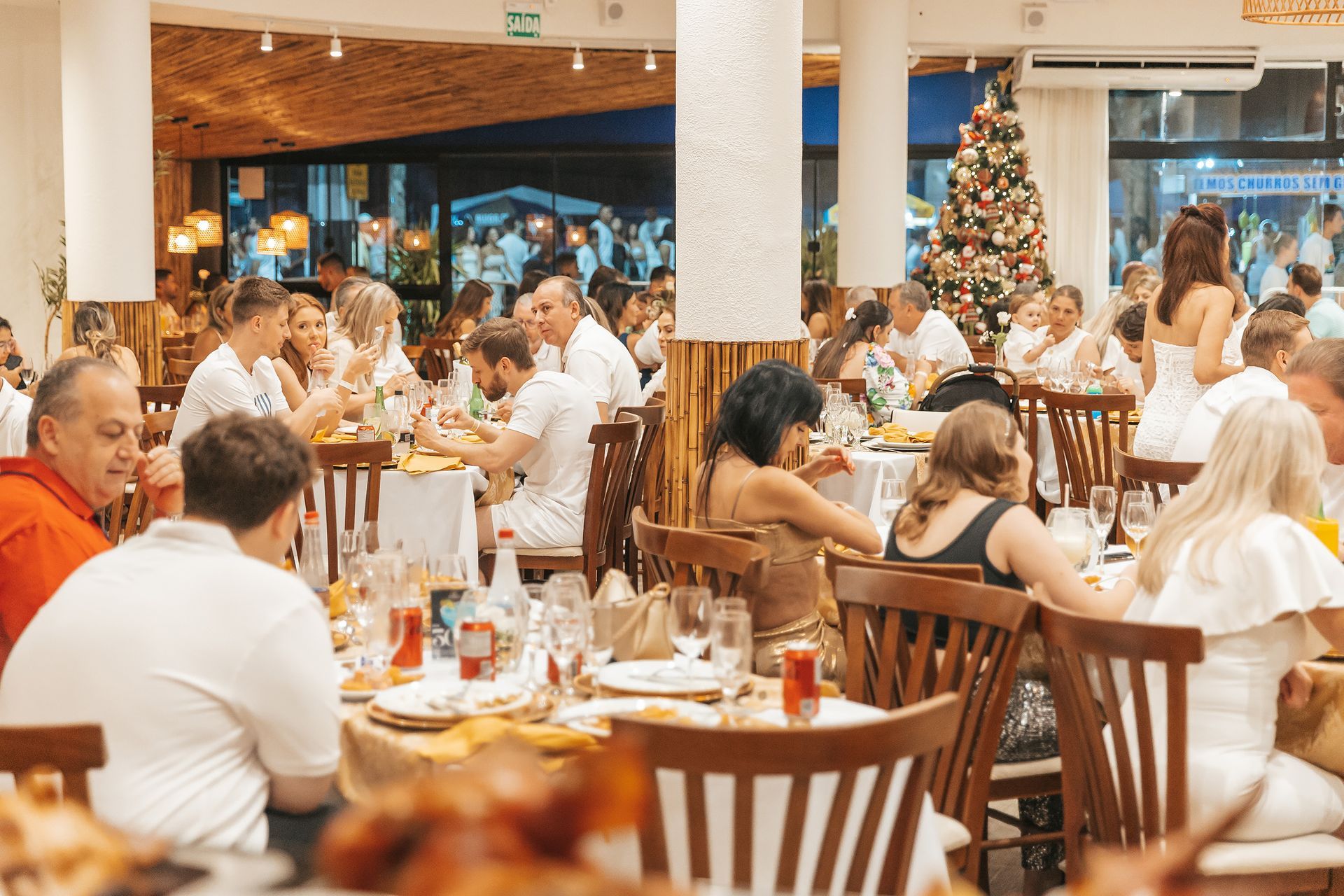 Sala de jantar festiva de um restaurante com muitos clientes sentados às mesas e uma árvore de Natal decorada ao fundo.