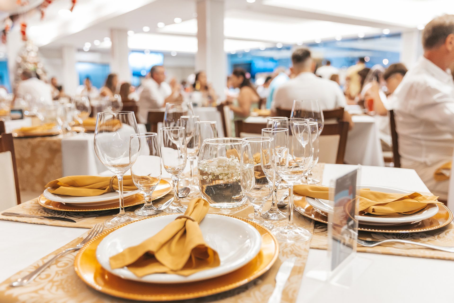 Elegante mesa de banquete posta com guardanapos dourados, taças de vinho e pratos em um salão de restaurante iluminado.