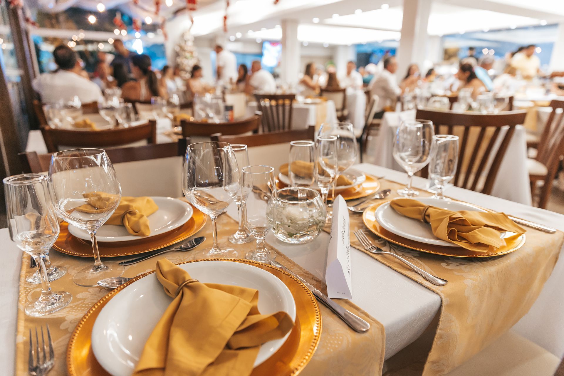 Elegante mesa de banquete com guardanapos dourados, taças de cristal e toalha de mesa branca em um salão de eventos iluminado.