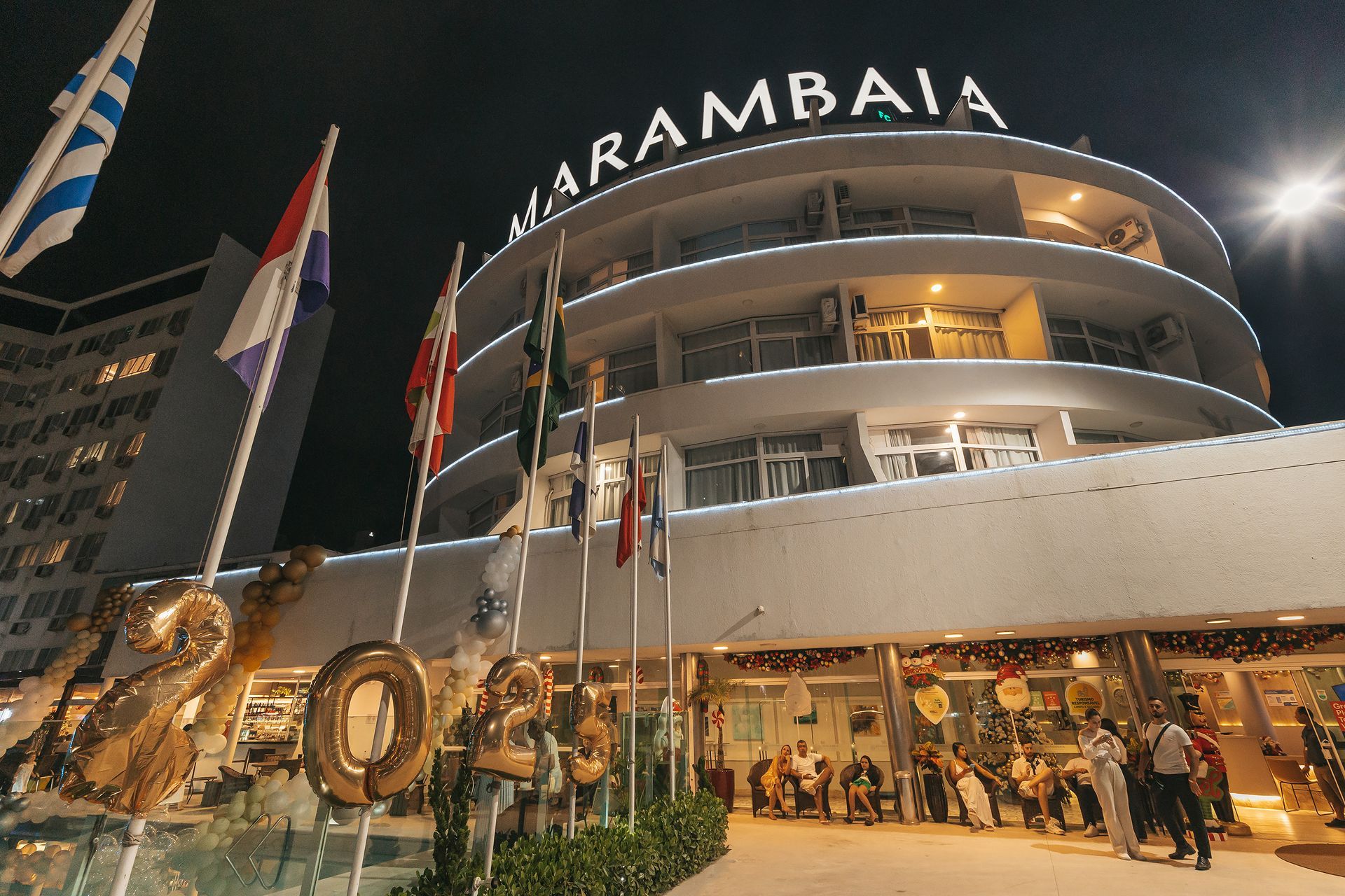 Vista noturna do edifício Karambai com varandas iluminadas, bandeiras e pessoas reunidas em frente à entrada.
