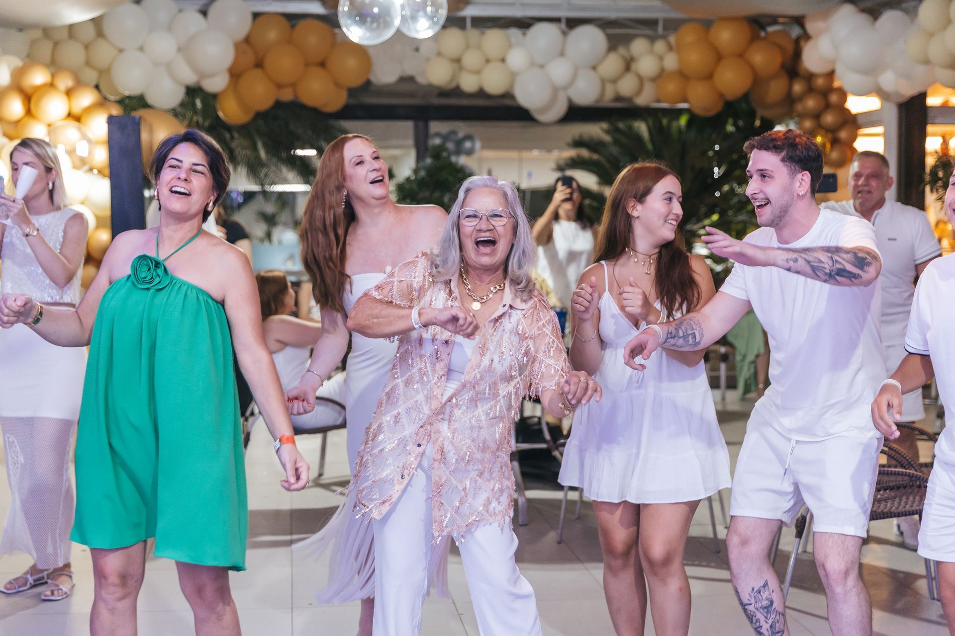 Grupo dançando e sorrindo em uma festa festiva em ambiente fechado com balões brancos e dourados.