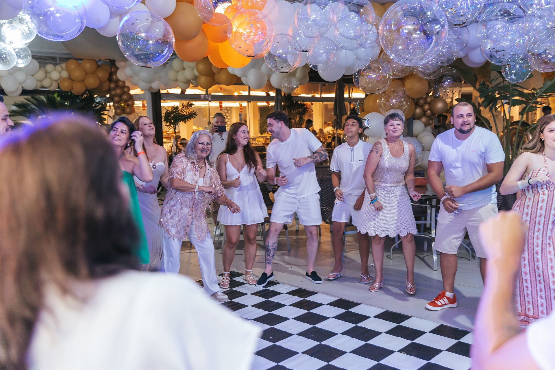Pessoas dançando em uma festa animada em ambiente fechado, sob decorações de balões e luzes aconchegantes.