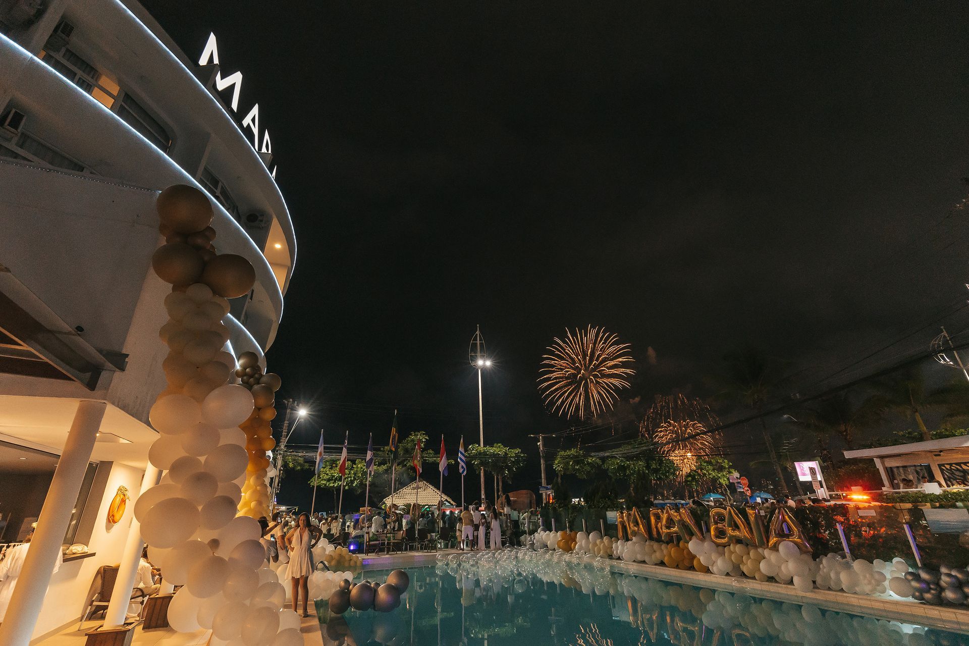 Espetáculo noturno de fogos de artifício à beira da piscina, ao lado de um prédio iluminado do resort e palmeiras.