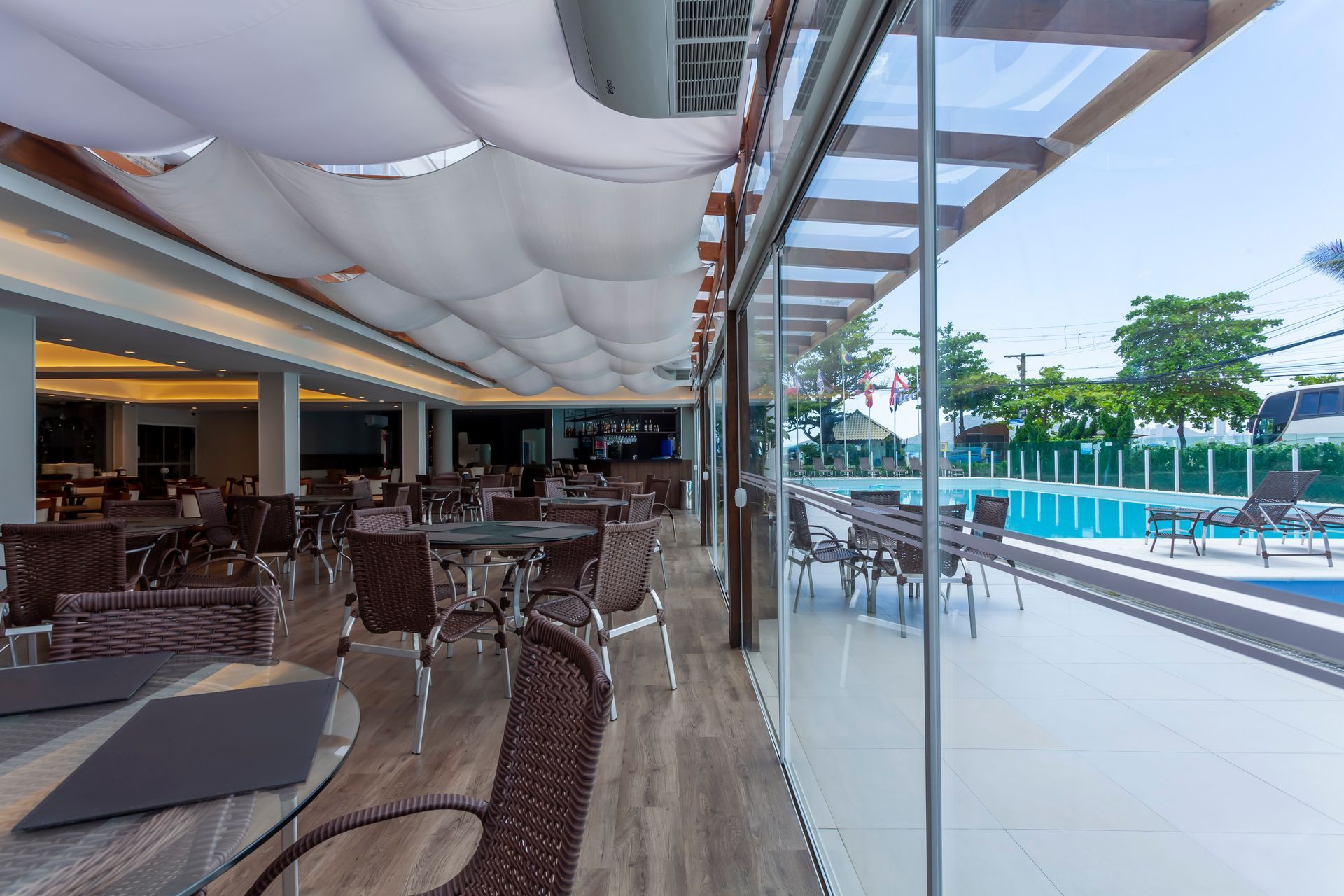 Área de refeições ao ar livre no resort, com cadeiras e mesas de vime ao lado da piscina, sob um toldo que proporciona sombra.