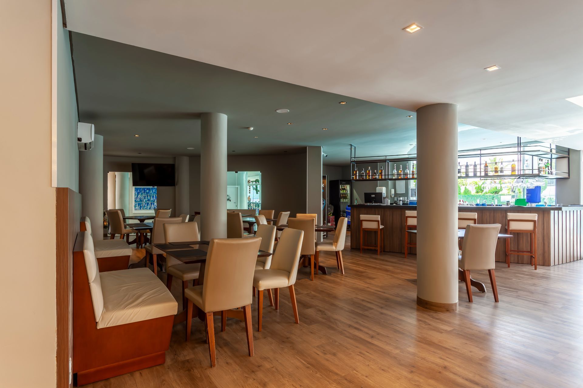 Ampla e moderna área de jantar do restaurante com piso de madeira, cadeiras bege e um balcão de bar ao fundo.