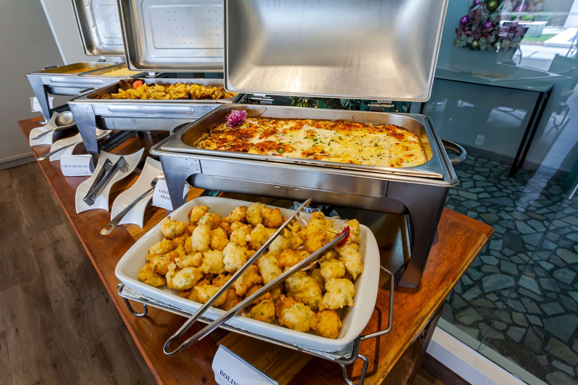 Mesa de bufê com bandejas de pratos assados ​​e pegadores, incluindo caçarola e bolinhos de batata.