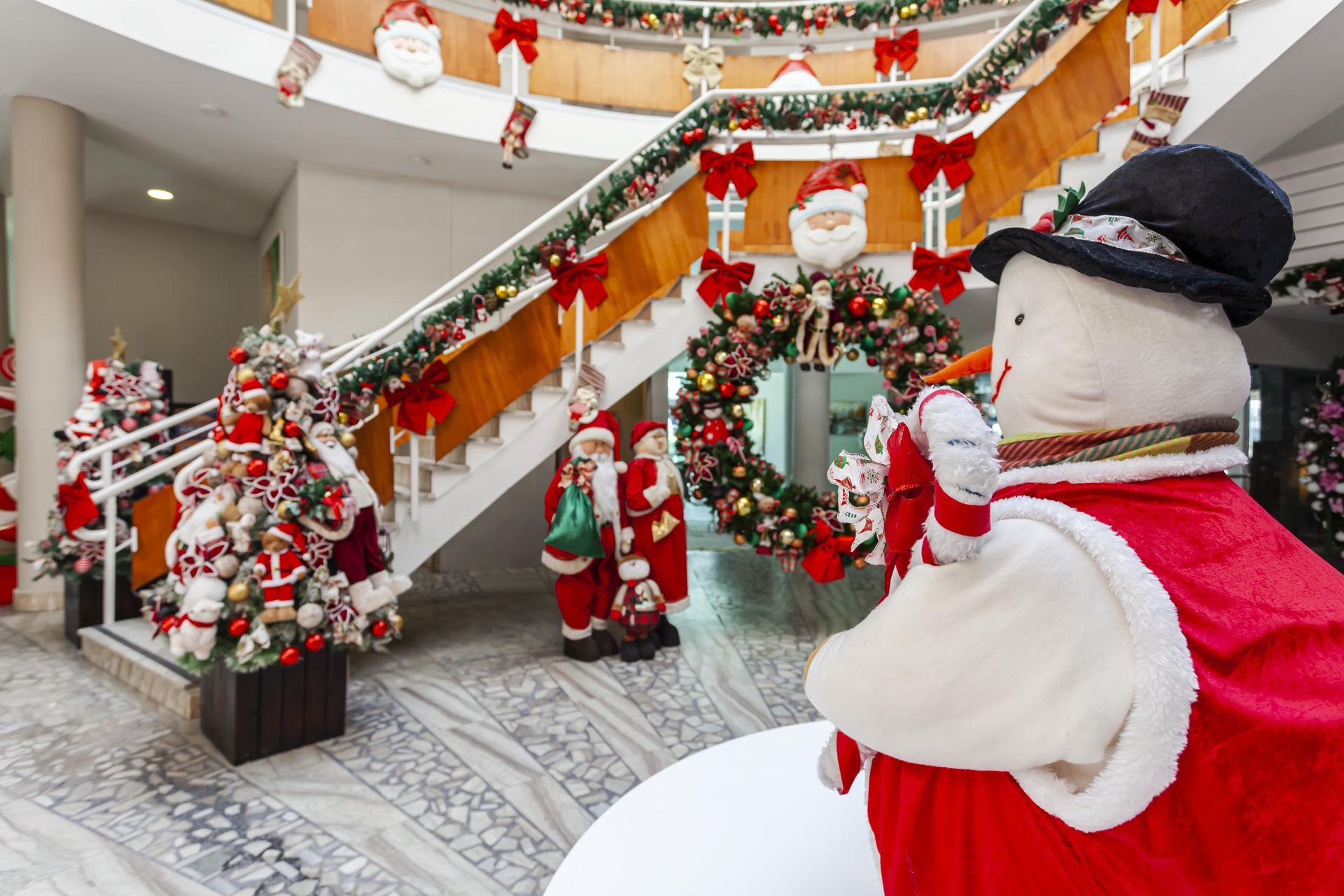 Decoração natalina festiva em ambiente interno com figuras do Papai Noel, guirlandas, grinaldas e um boneco de neve decorativo junto a uma escada.