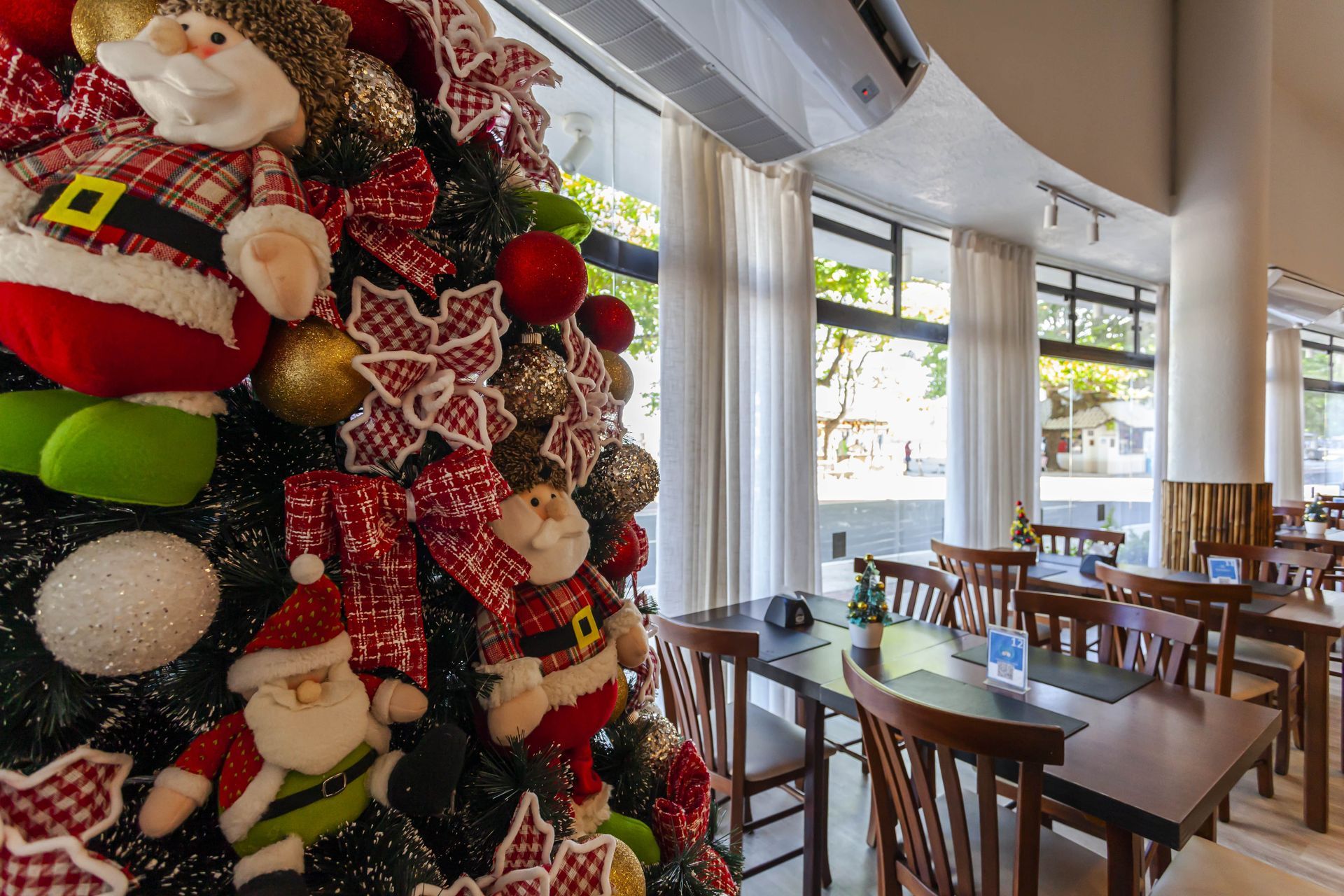Árvore de Natal festiva com enfeites de Papai Noel ao lado de uma área de estar de café bem iluminada.