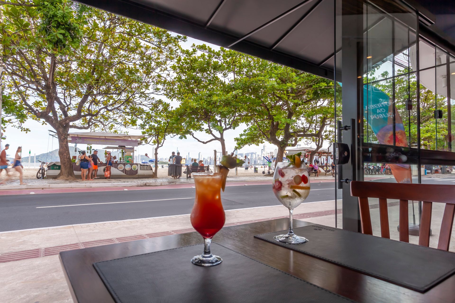 Coquetéis em uma mesa de café com vista para um calçadão arborizado à beira-mar.