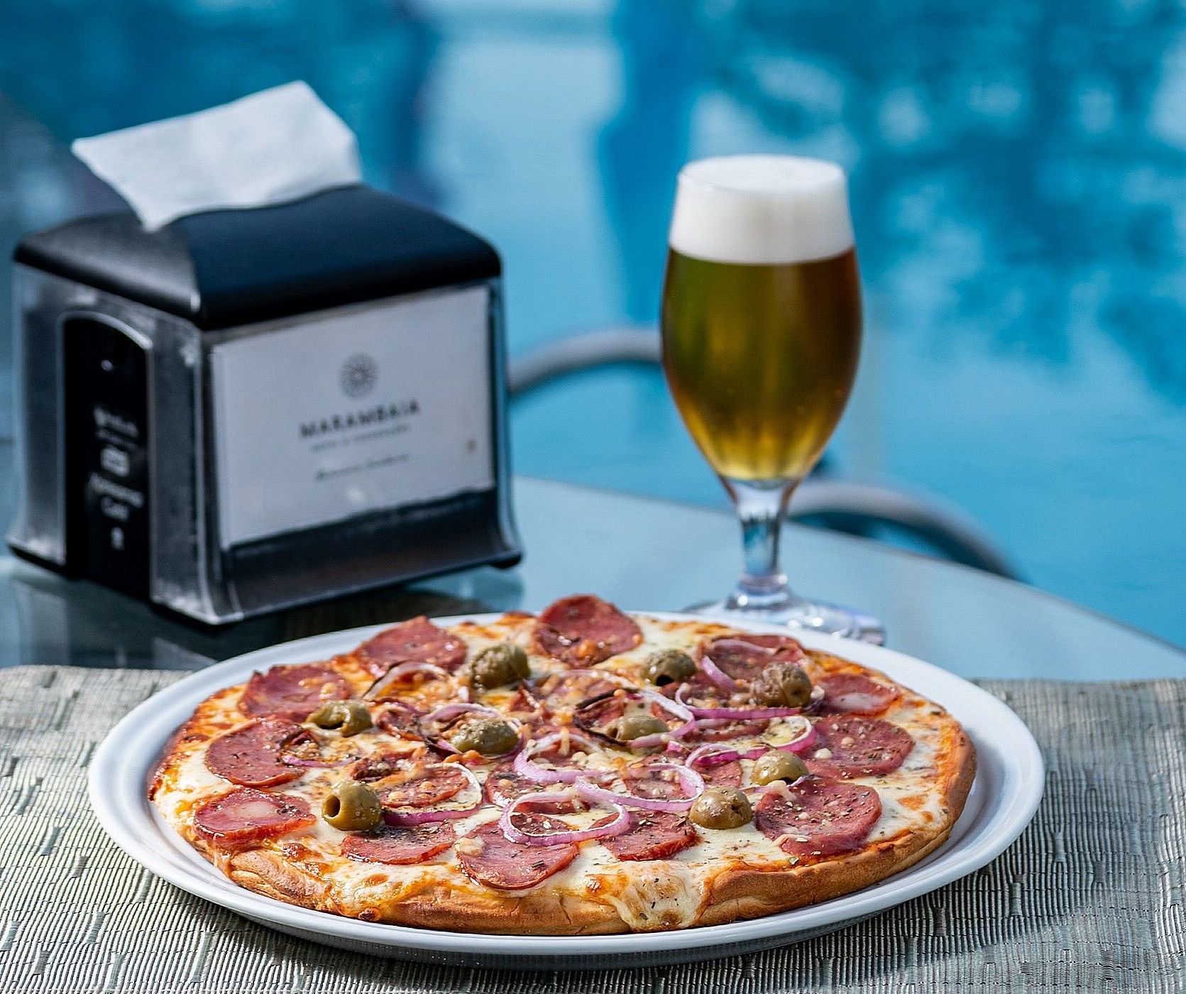 Pizza de pepperoni em uma mesa ao lado de um copo de cerveja e um dispensador de guardanapos perto da piscina.