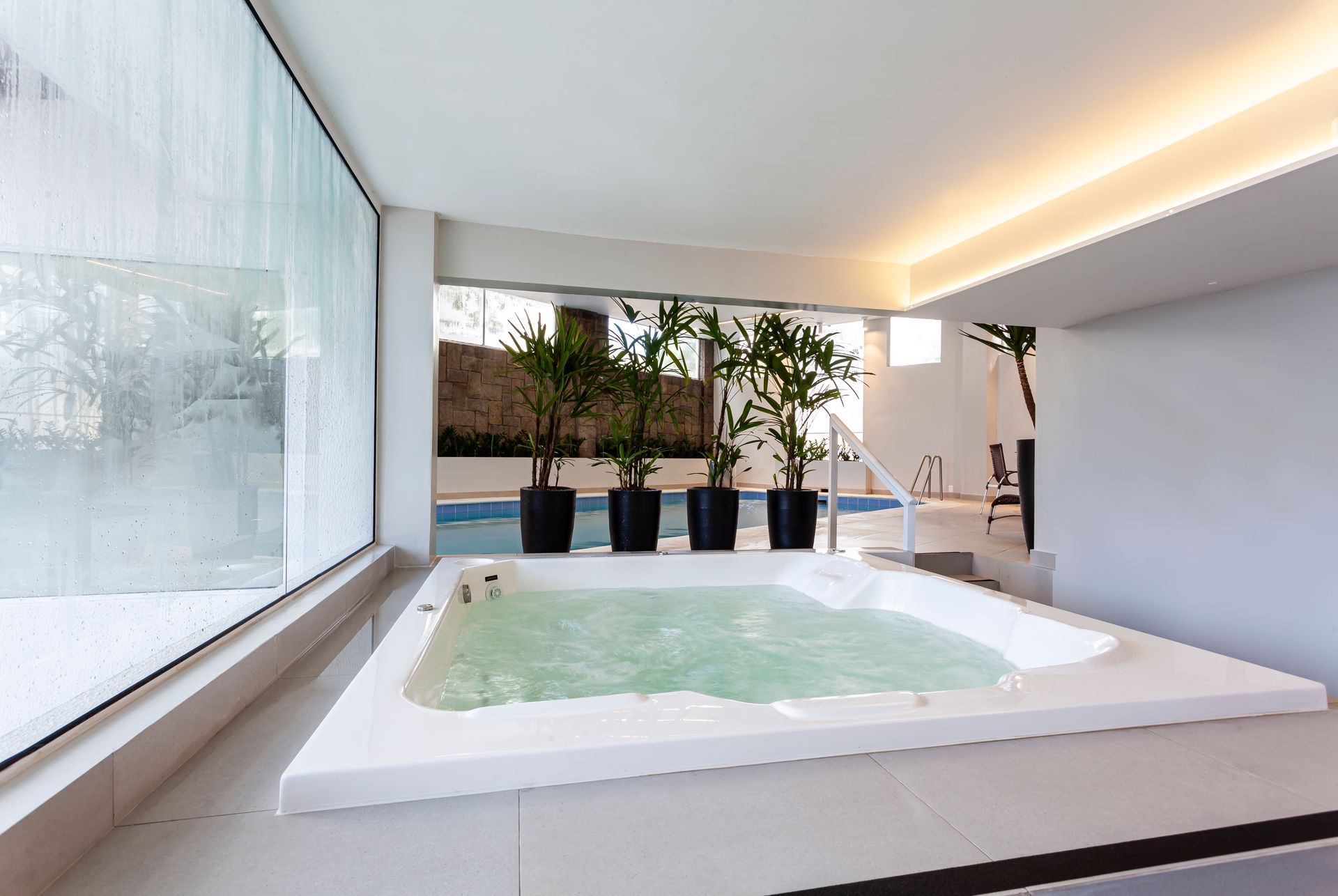 Spa moderno com banheira de hidromassagem, plantas em vasos e grandes janelas em um ambiente minimalista e luminoso.