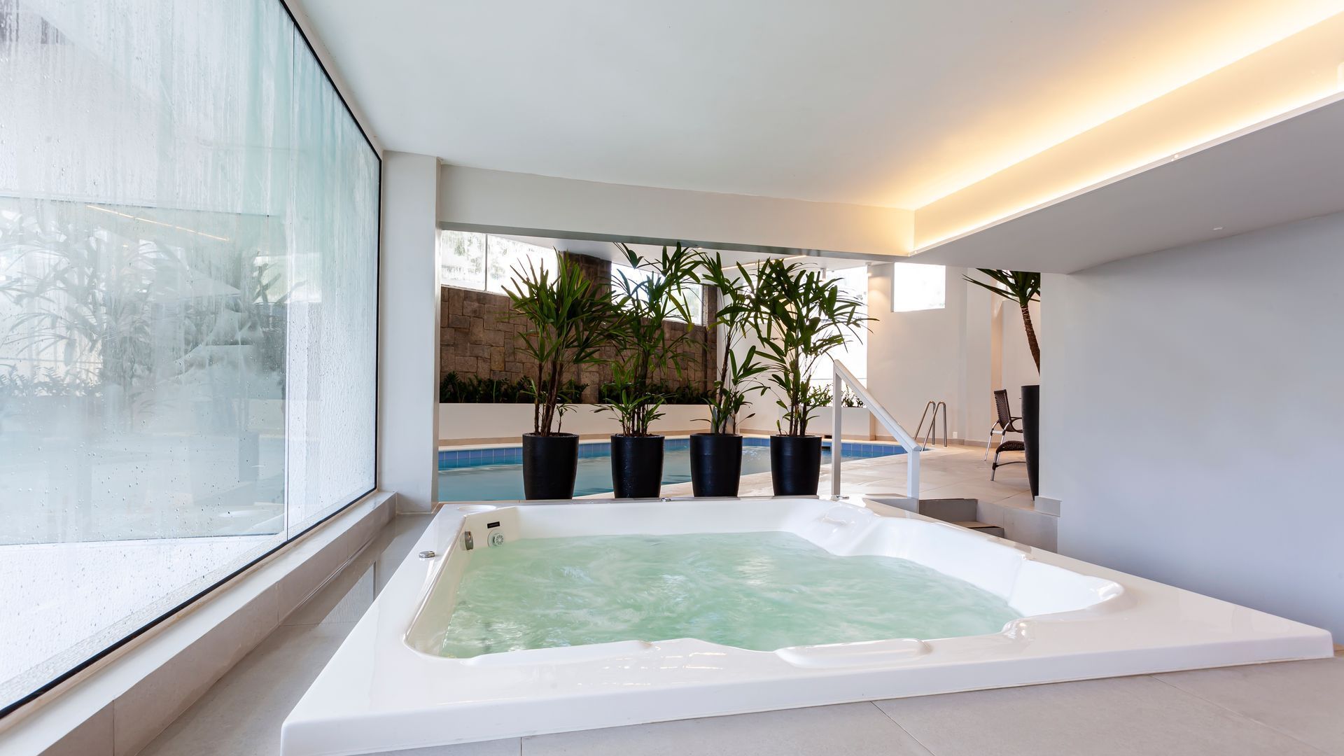 Piscina de spa interna moderna com paredes brancas, grandes janelas e plantas em vasos ao fundo.