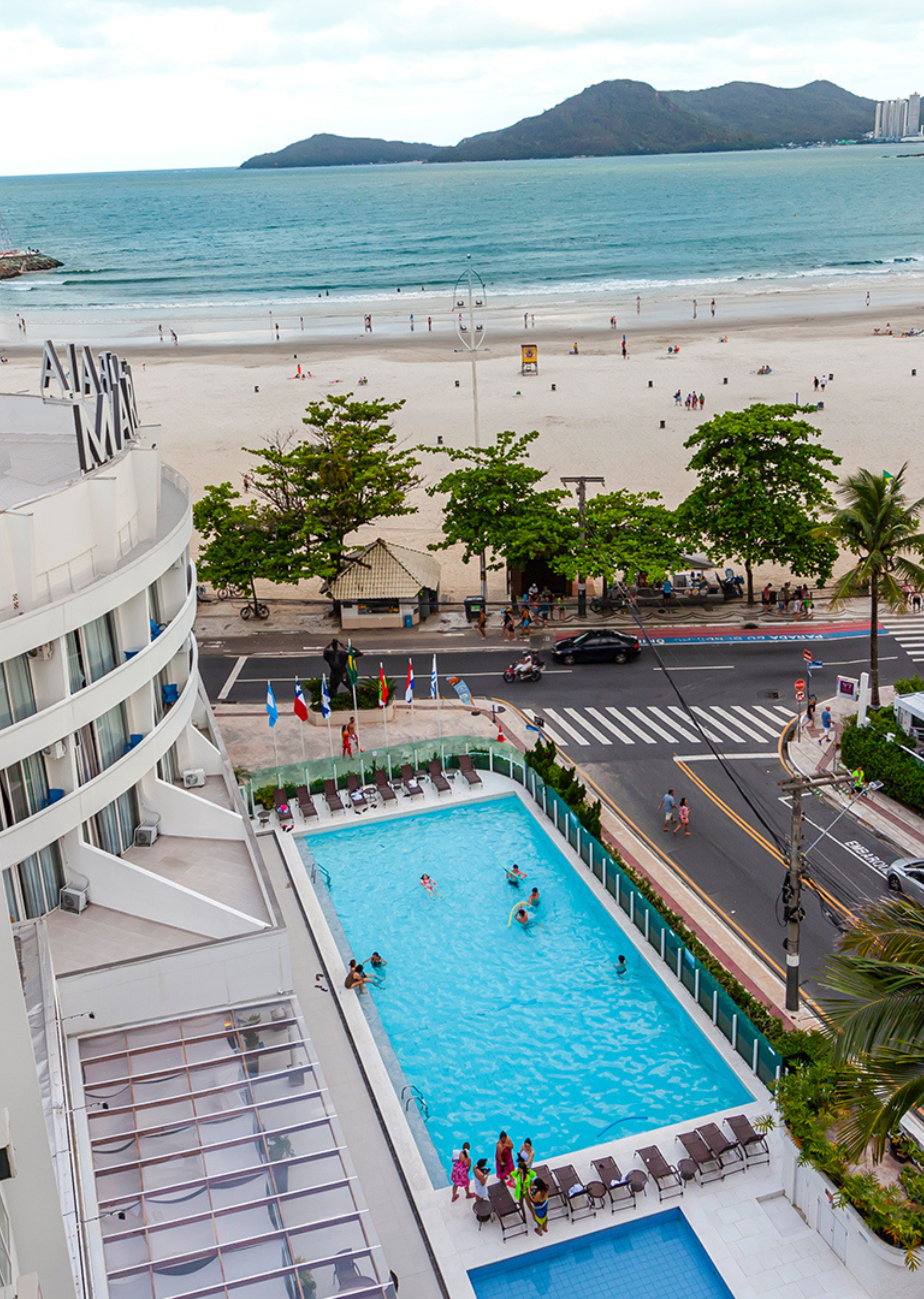 Piscina de hotel à beira-mar com vista para uma praia de areia, estrada e mar turquesa com ilhas ao longe.