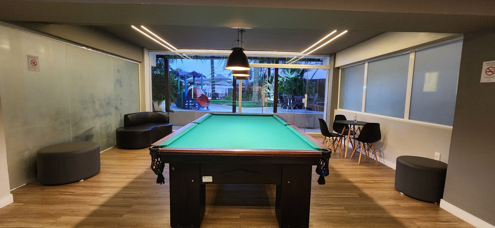 Sala de jogos moderna com mesa de bilhar, piso de madeira, sofás e grandes janelas com vista para a água.