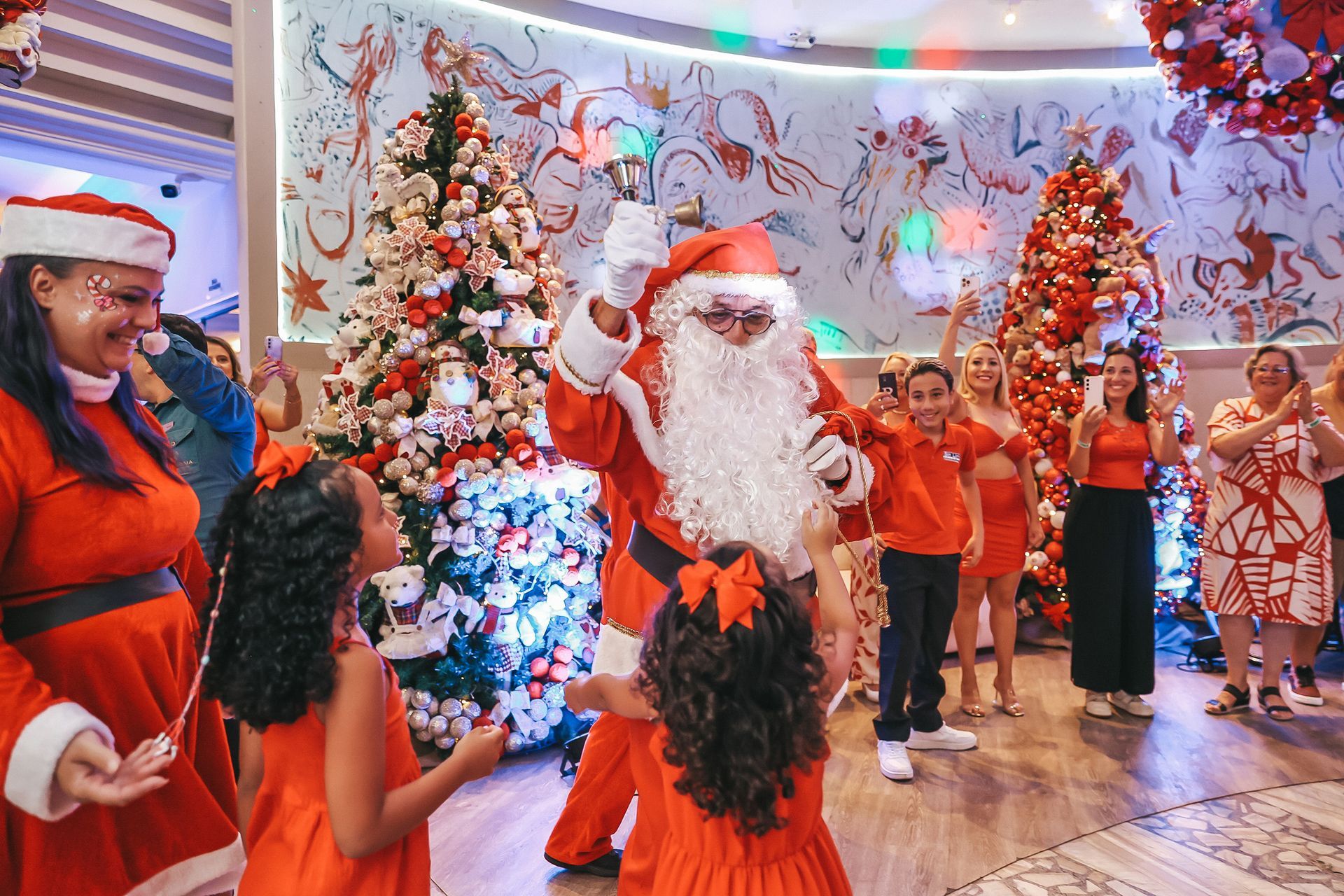 Papai Noel e crianças dançando em trajes festivos vermelhos ao lado de árvores de Natal decoradas.