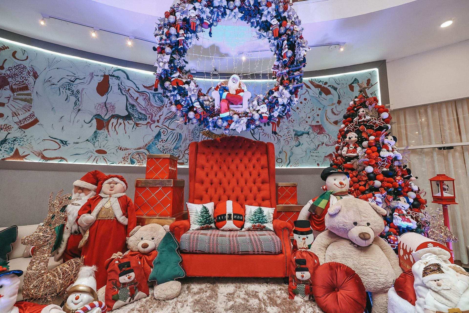 Decoração natalina com Papai Noel, ursinhos de pelúcia, cadeira vermelha e árvore decorada em um ambiente festivo.