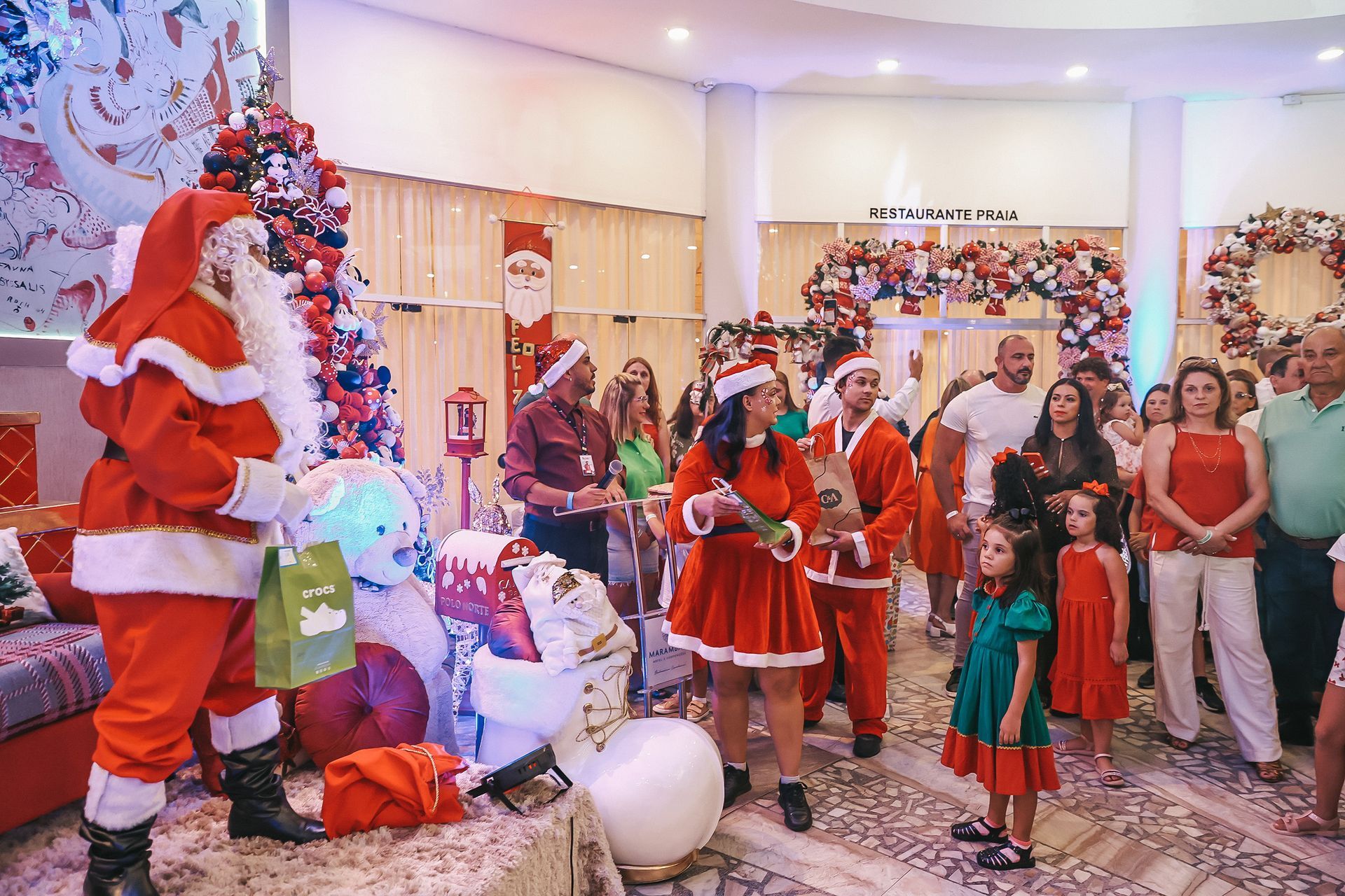 Papai Noel e crianças celebrando o Natal em uma sala decorada com uma árvore iluminada e presentes.