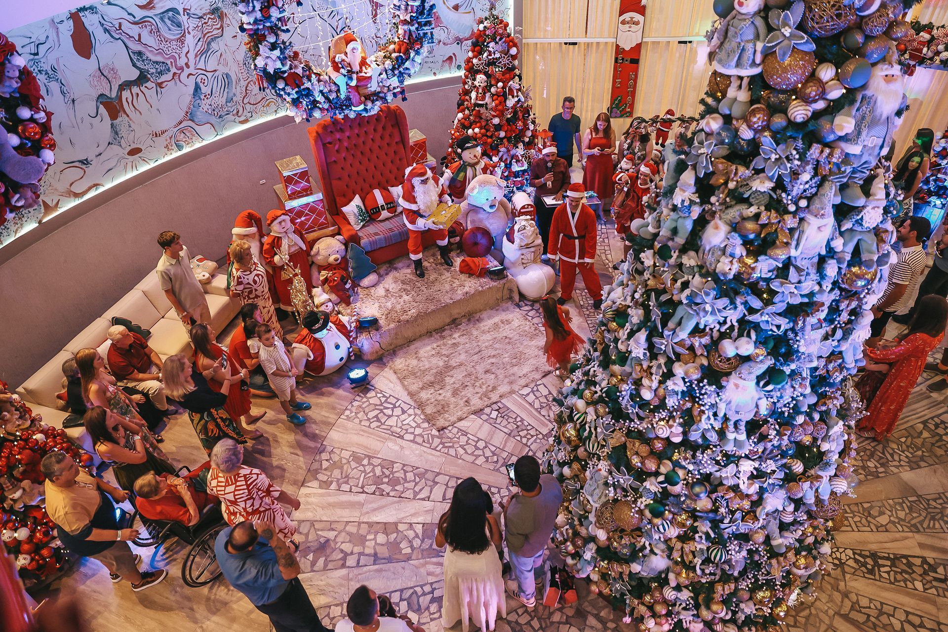 Decoração festiva em ambiente interno com uma grande árvore de Natal decorada e crianças reunidas em torno de uma estrutura semelhante a um carrossel.