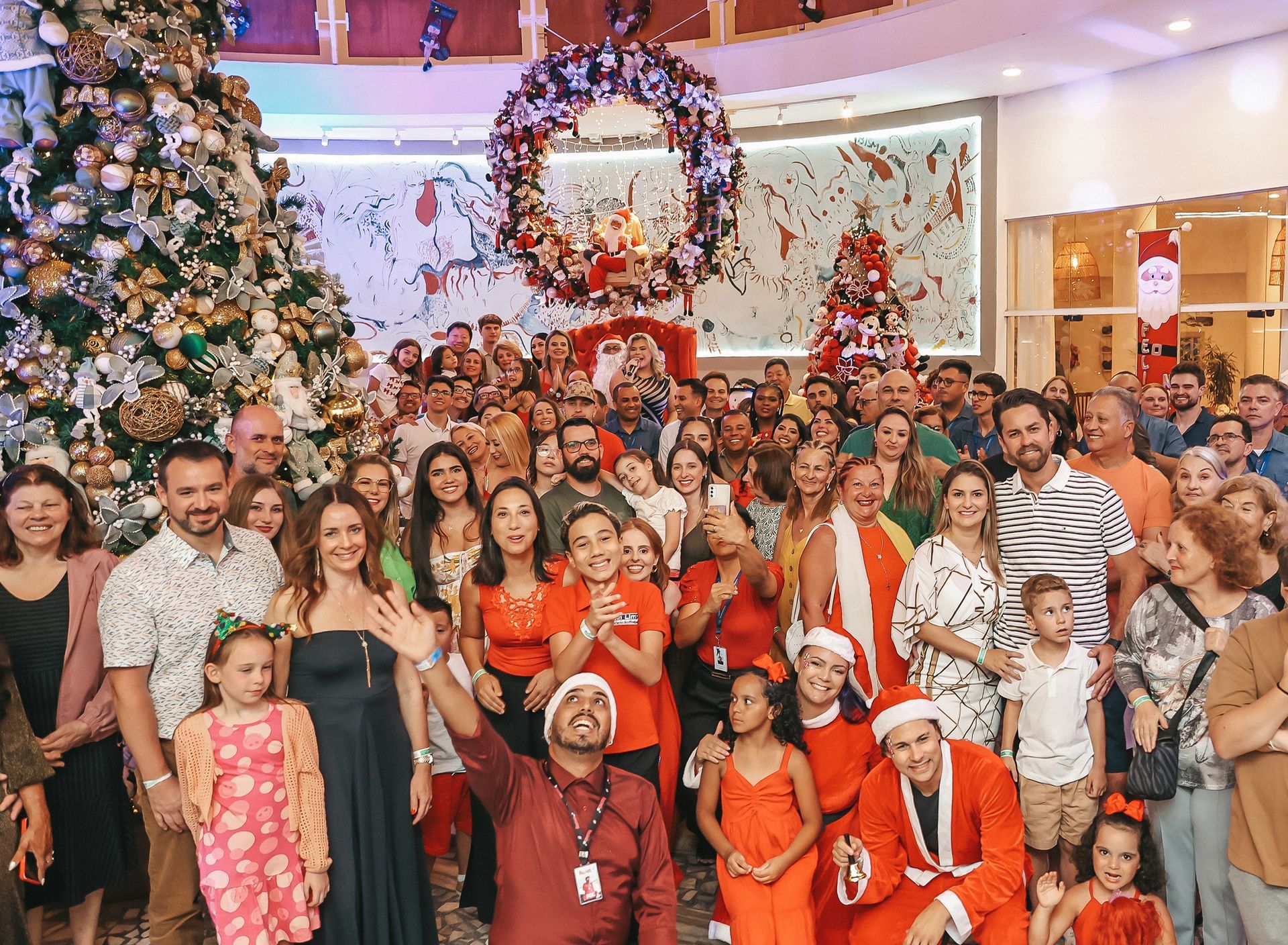 Grande grupo de amigos posando em frente a uma árvore de Natal e guirlanda decoradas, muitos com trajes festivos vermelhos.