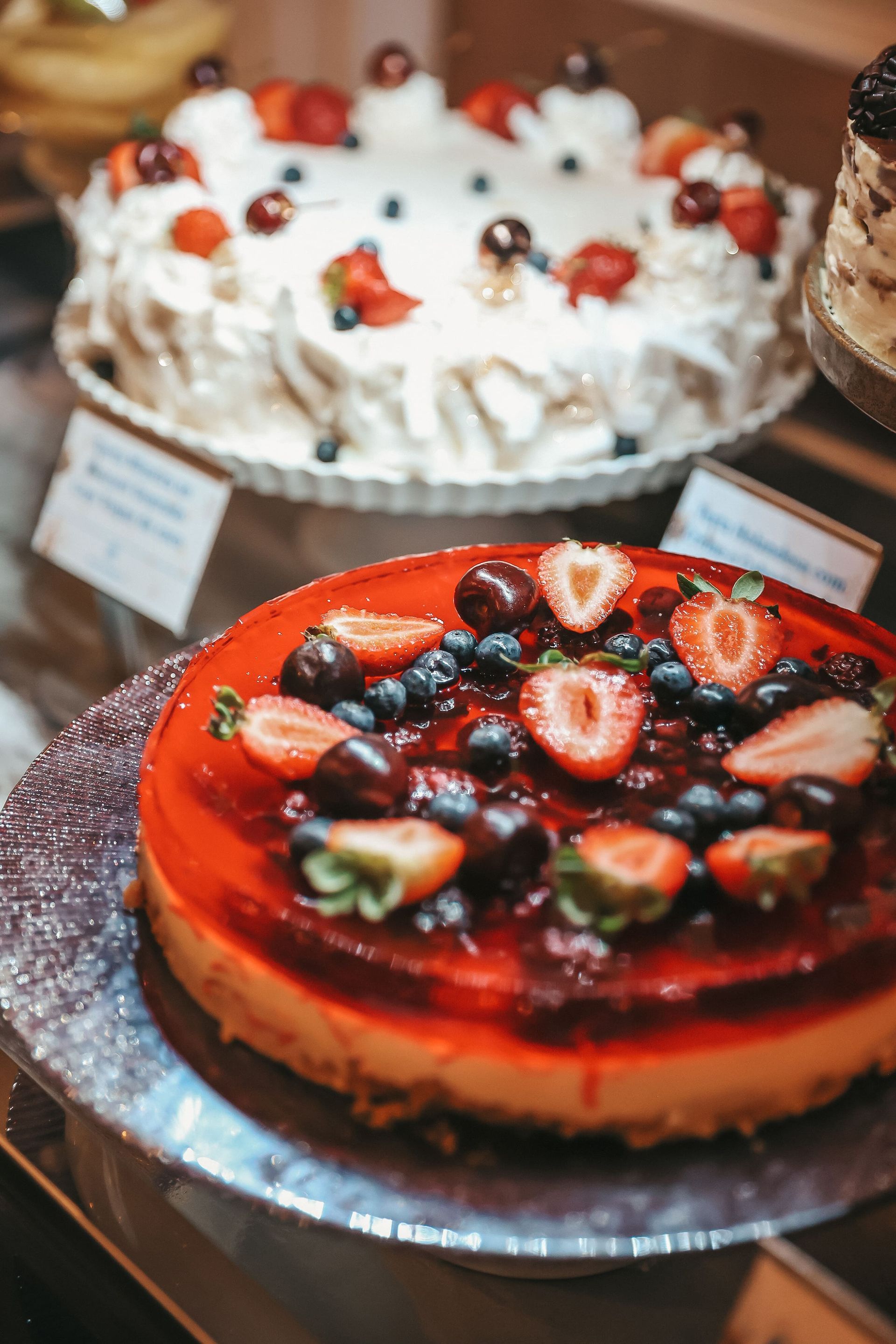 Em exposição, dois bolos decorados: um bolo vermelho com cobertura de frutas e, atrás dele, um bolo branco de morango.