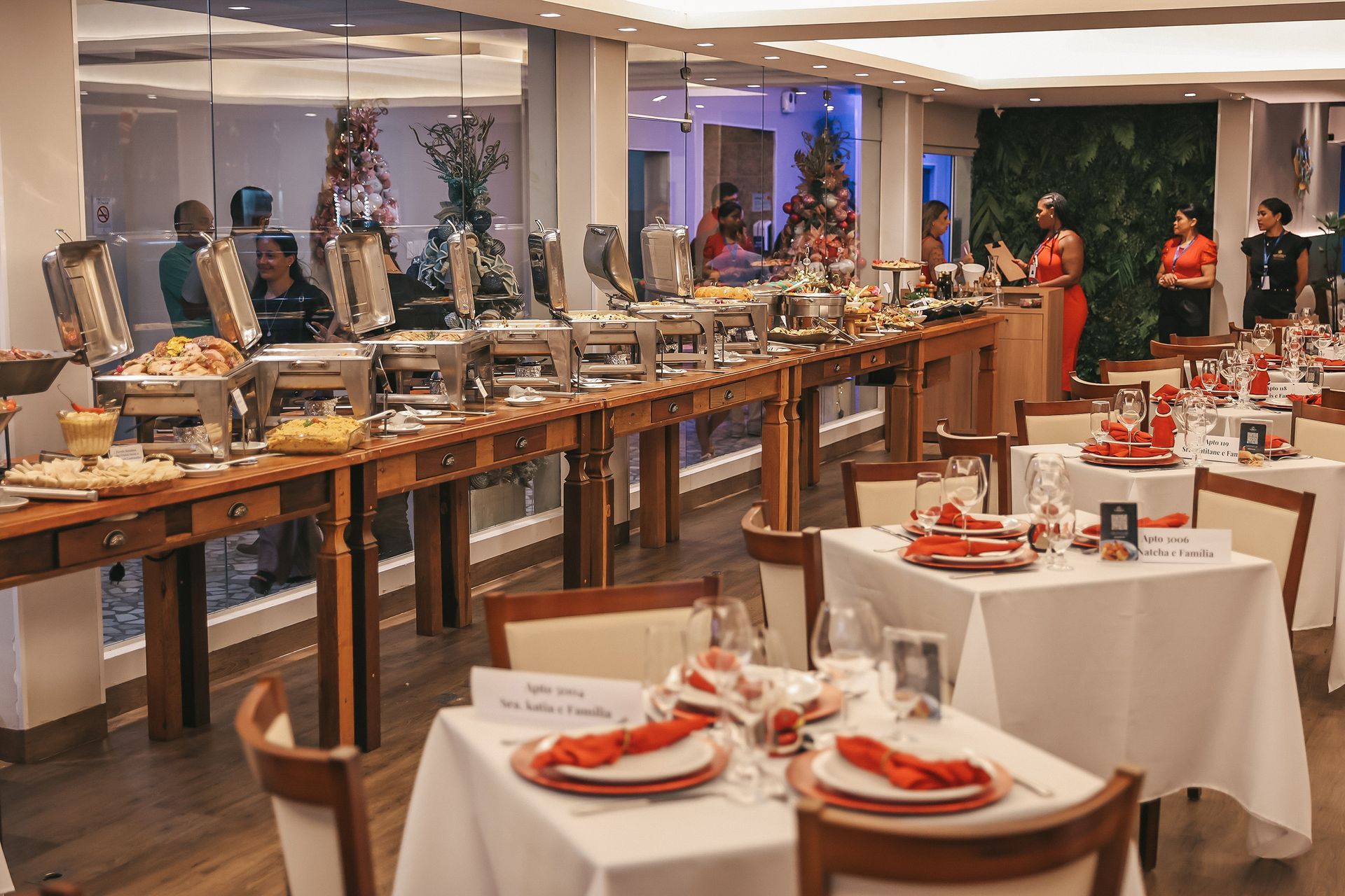 Elegante buffet montado em um salão de banquetes com toalhas de mesa brancas, lugares marcados em vermelho e convidados ao fundo.