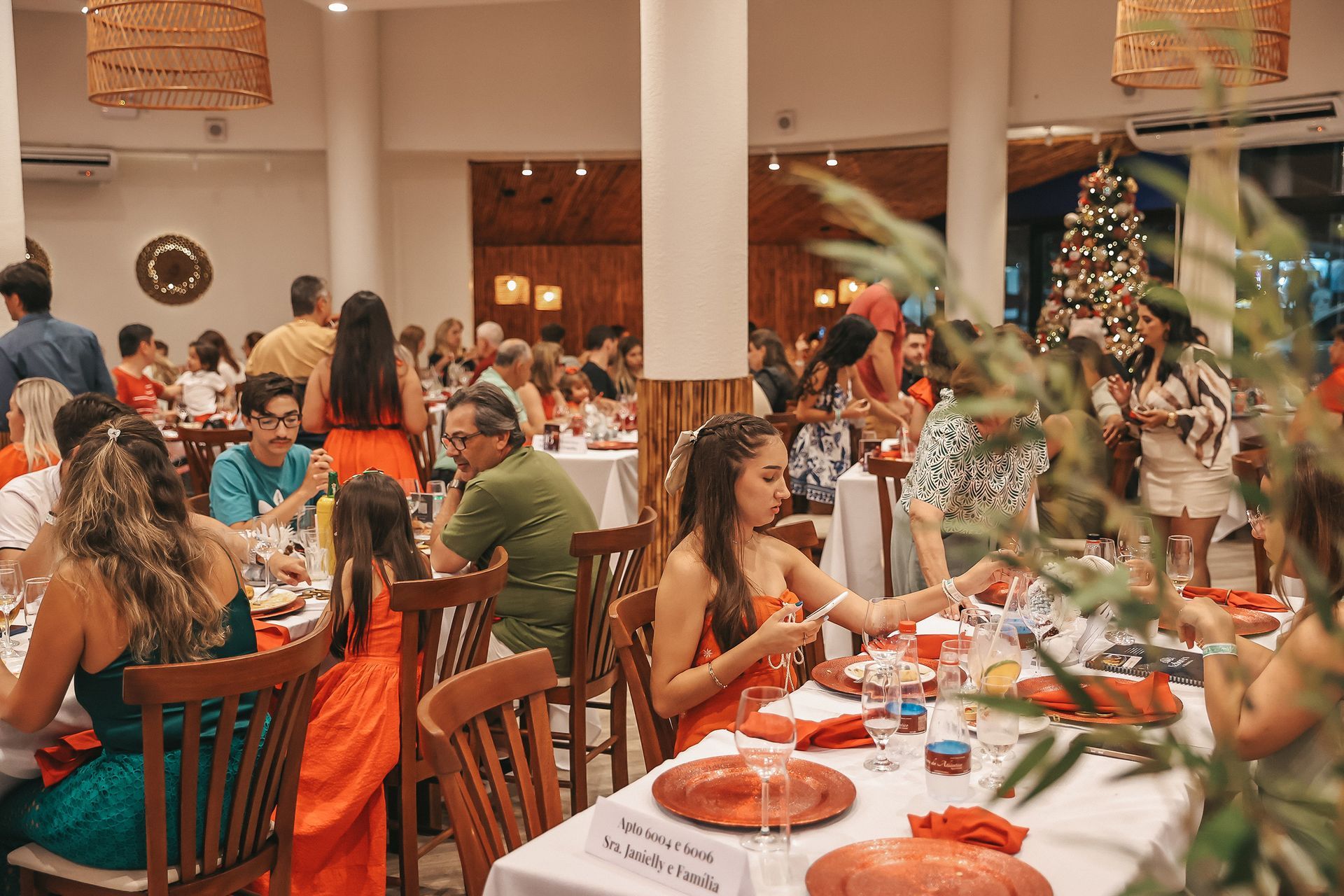 Pessoas jantando em um restaurante festivo com mesas postas em vermelho e decorações natalinas ao fundo.
