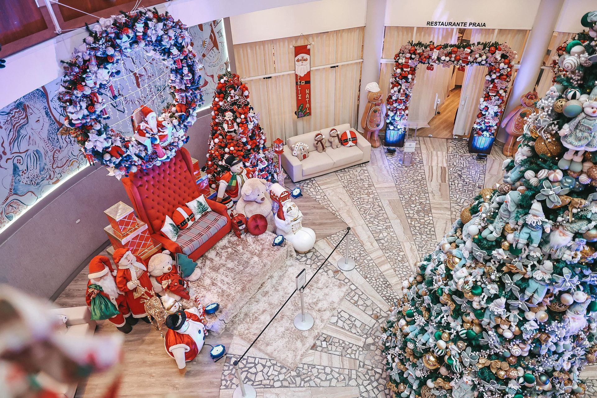 Sala de estar festiva com árvores de Natal decoradas, guirlandas, presentes e uma escadaria com enfeites natalinos.