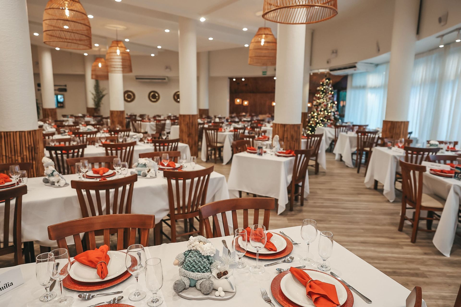 Sala de jantar de restaurante iluminada com toalhas de mesa brancas, cadeiras de madeira e luminárias pendentes de vime.