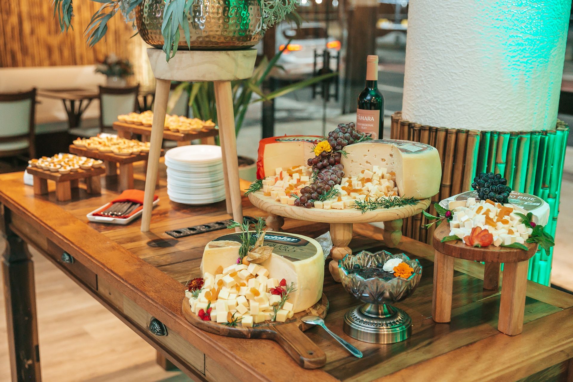 Mesa de bufê com queijos variados, biscoitos, frutas e pães em suportes de madeira.