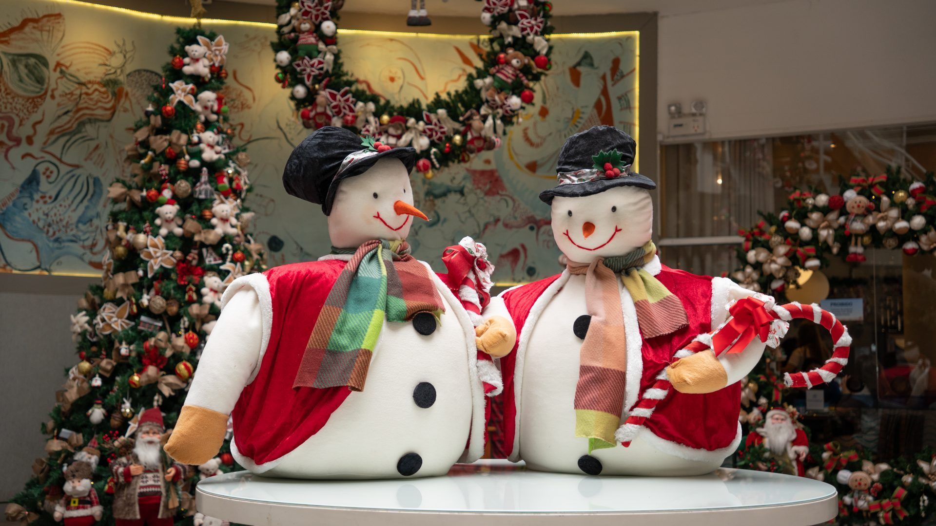 Duas decorações natalinas em forma de boneco de neve sobre uma mesa, com árvores de Natal e guirlandas ao fundo.