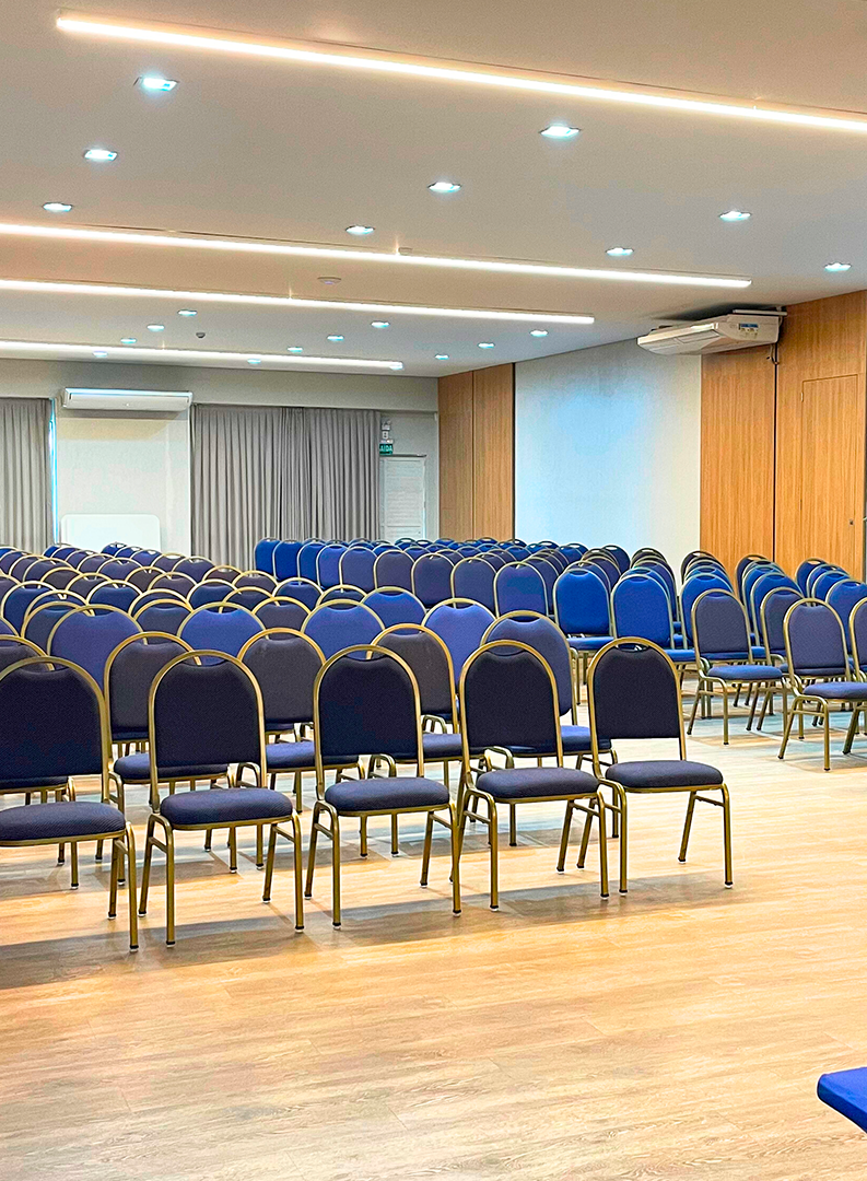 Sala de conferências vazia com fileiras de cadeiras azuis voltadas para um palco frontal iluminado por luzes no teto.
