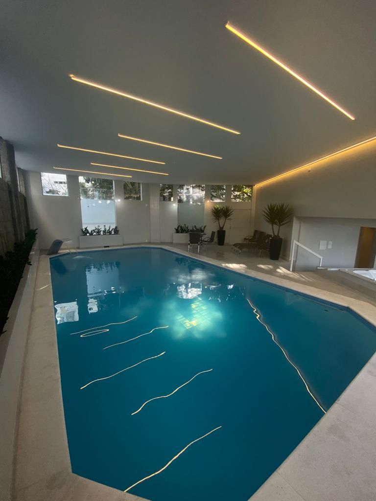 Piscina coberta com água azul, paredes brancas e longas luminárias de teto que se refletem na superfície.