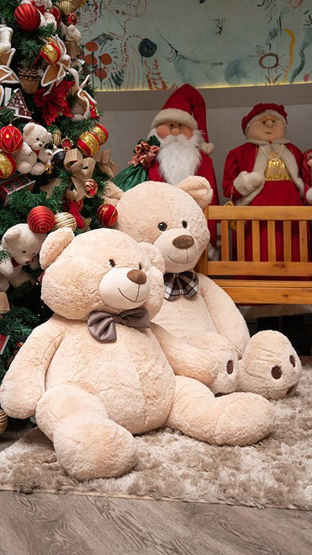 Três ursinhos de pelúcia bege em uma decoração natalina ao lado de uma árvore de Natal decorada e figuras do Papai Noel.