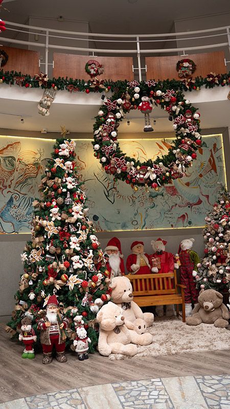 Decoração natalina festiva com árvores de Natal decoradas, guirlandas, ursinhos de pelúcia e arranjos florais no saguão de um shopping.