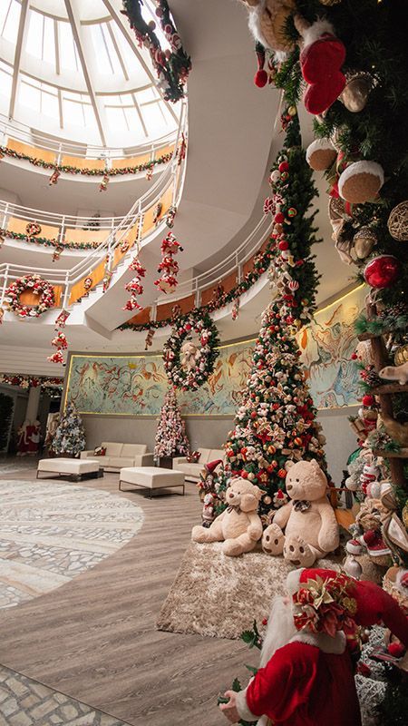 Hall de entrada festivo com guirlandas natalinas, ursinhos de pelúcia e enfeites vermelhos e dourados sob um teto com claraboia.