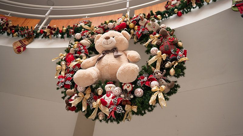 Um grande urso de pelúcia em uma guirlanda festiva de Natal com enfeites vermelhos, dourados e brancos em um saguão interno.