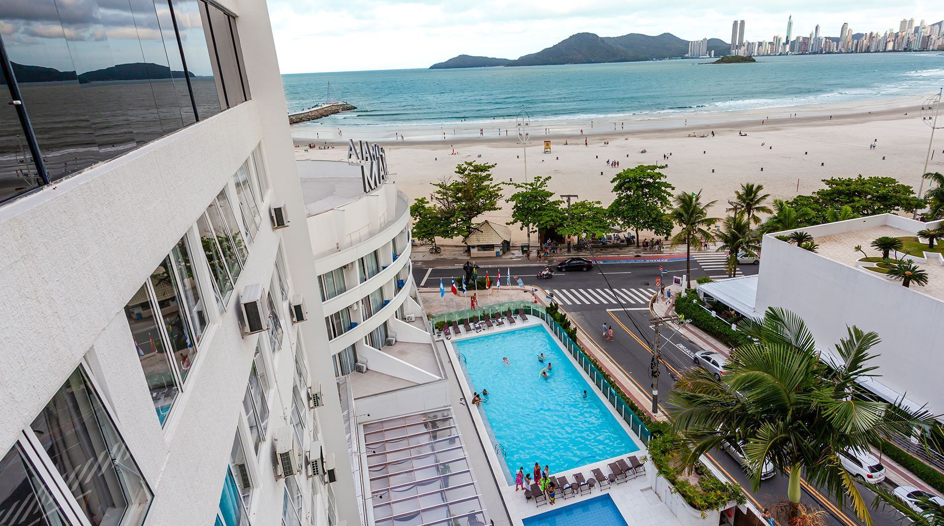 Piscina do hotel à beira-mar e vista para o oceano a partir da varanda, com o horizonte da cidade ao fundo.