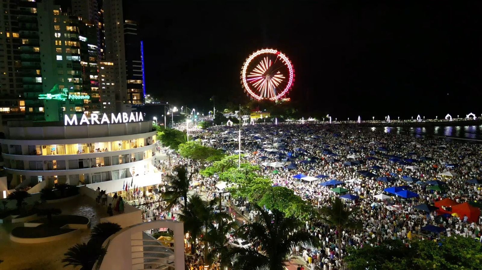 Concerto noturno lotado à beira-mar, com roda-gigante iluminada e letreiros de prédios iluminados.