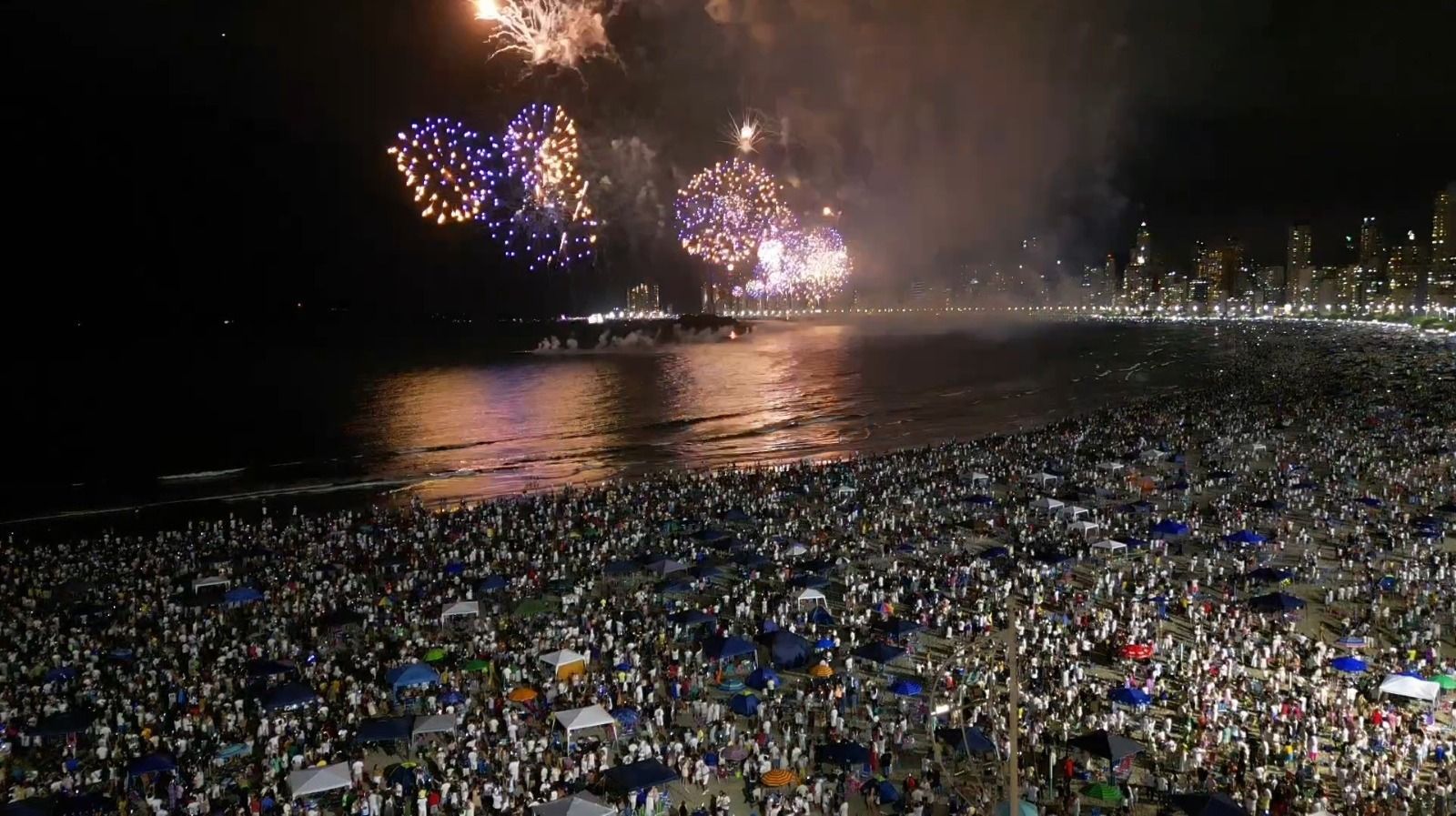 Praia lotada à noite, com fogos de artifício explodindo sobre a água e luzes da cidade ao longe.