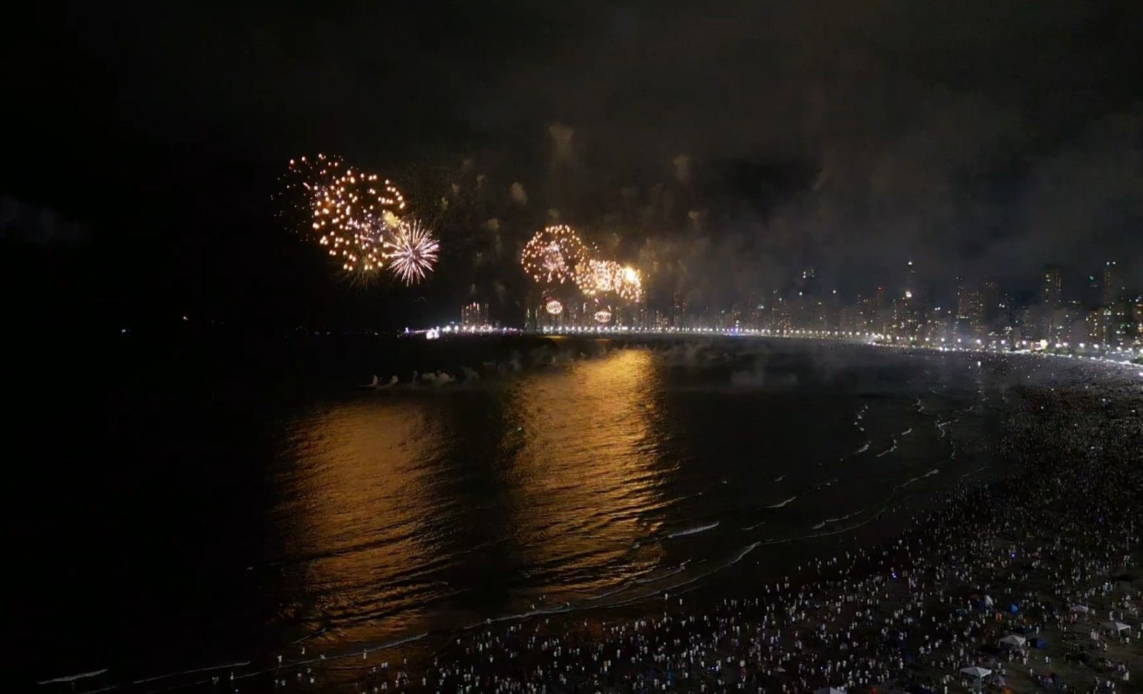 Fogos de artifício refletidos na escuridão da orla à noite, com luzes ao longo da costa.