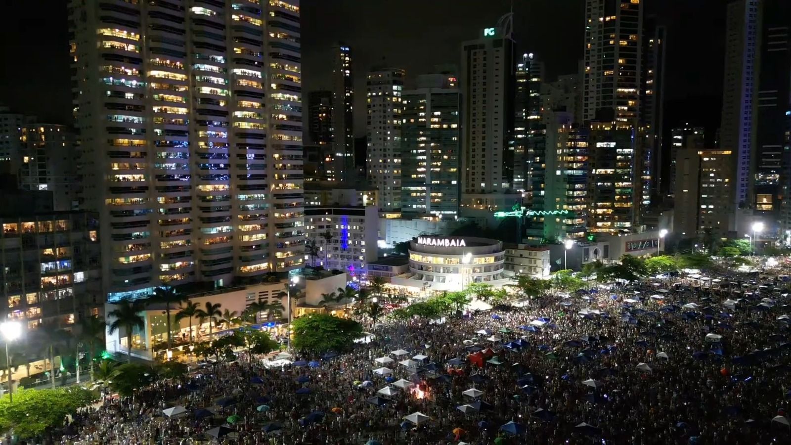 Praça da cidade à noite, repleta de pessoas, emoldurada por altos edifícios iluminados e luzes brilhantes.