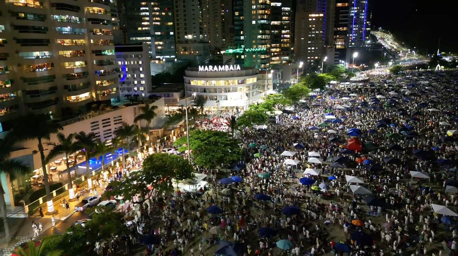 Praça da cidade à noite lotada de gente, cercada por arranha-céus iluminados e árvores.