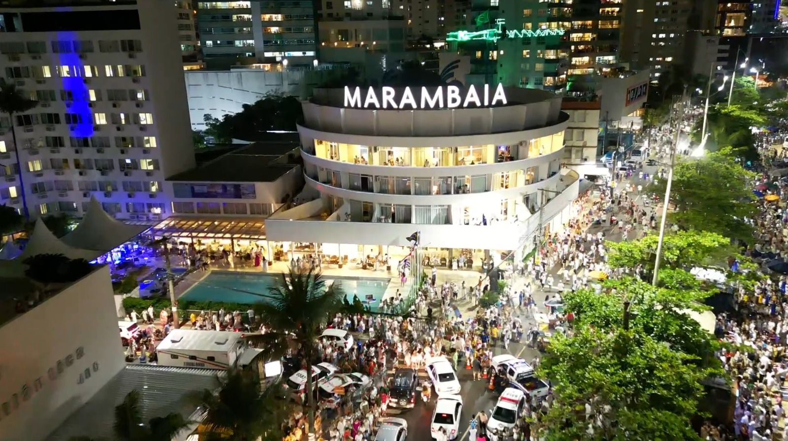 À noite, uma multidão se reúne em uma praça urbana iluminada, tendo o edifício MARAMBAIA ao centro.