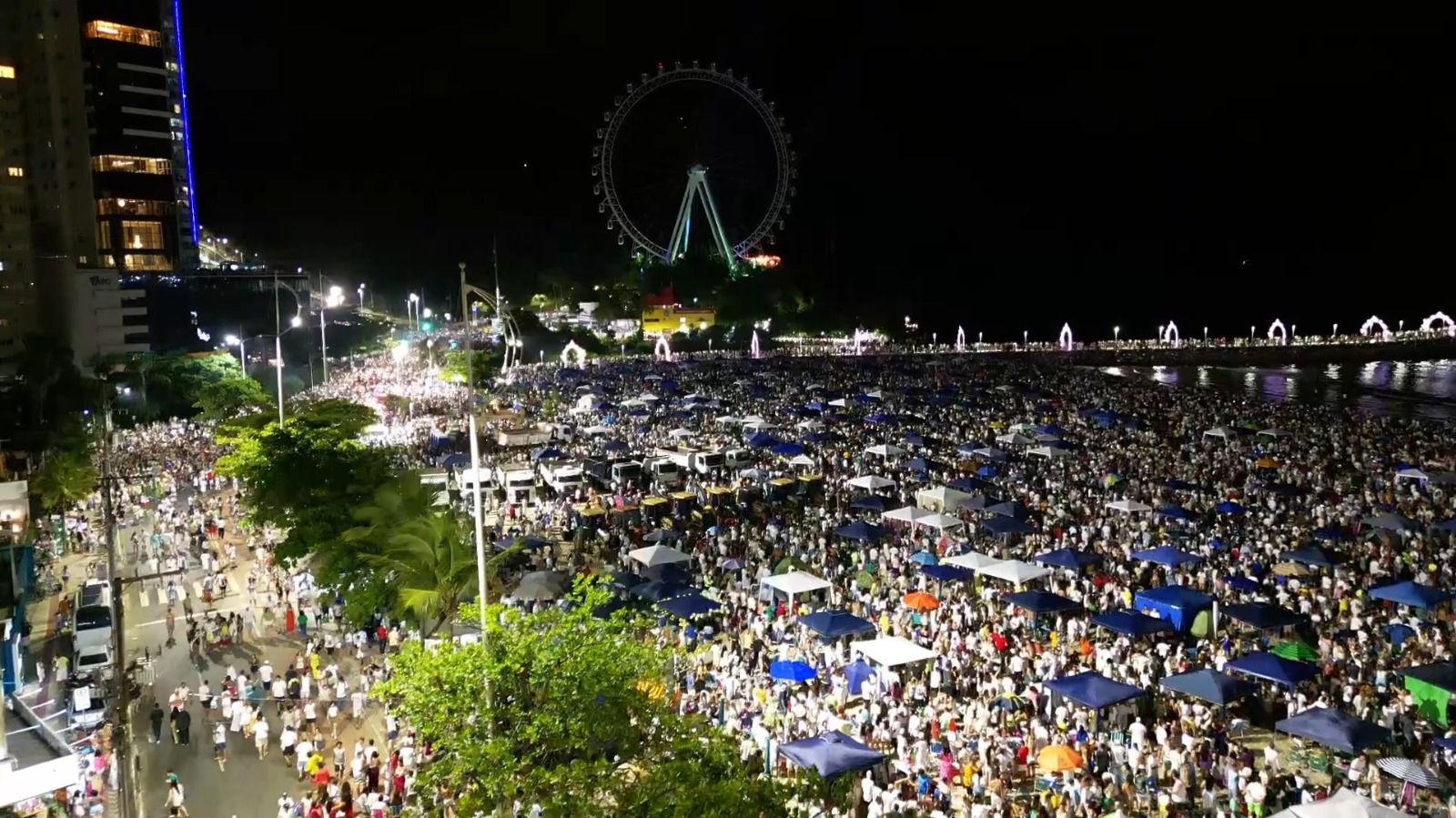 À noite, uma multidão lotava um espaço iluminado para festivais ao ar livre, com uma grande roda-gigante ao fundo.