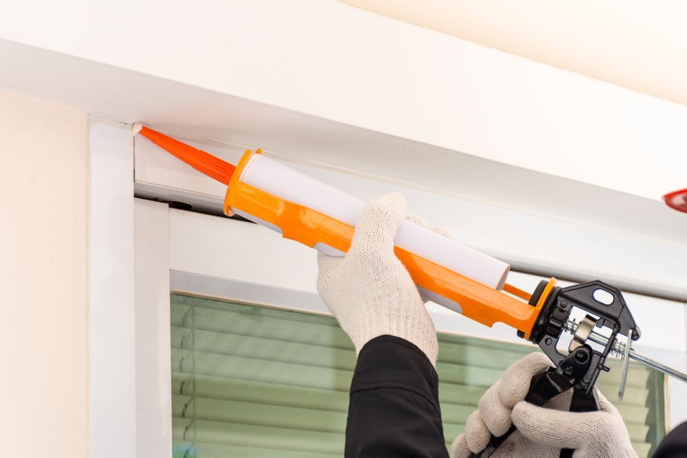 Gloved Hands Applying Orange Caulk With a Caulking Gun Around a Window Frame — C K Carpentry in Byron Bay, NSW