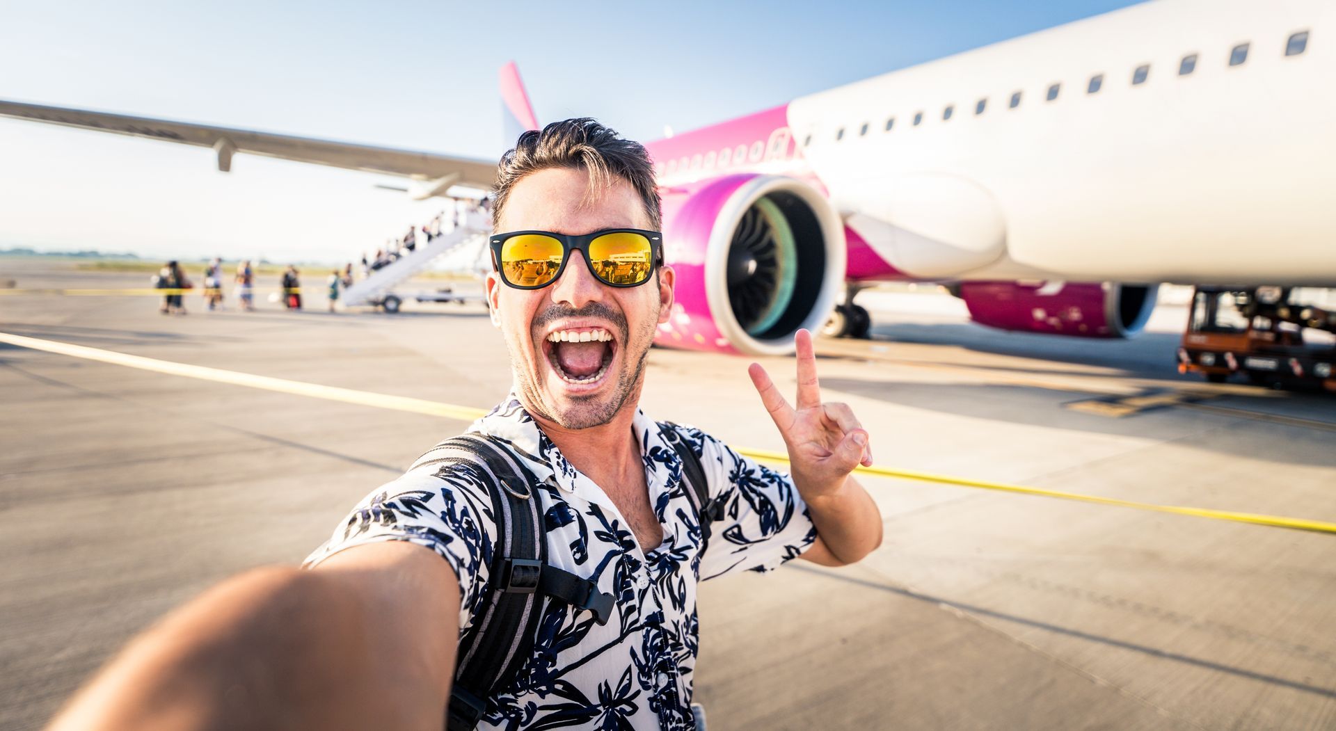 Man taking a selfie in front of a pink-and-white airplane on a sunny runway, smiling and flashing a peace sign