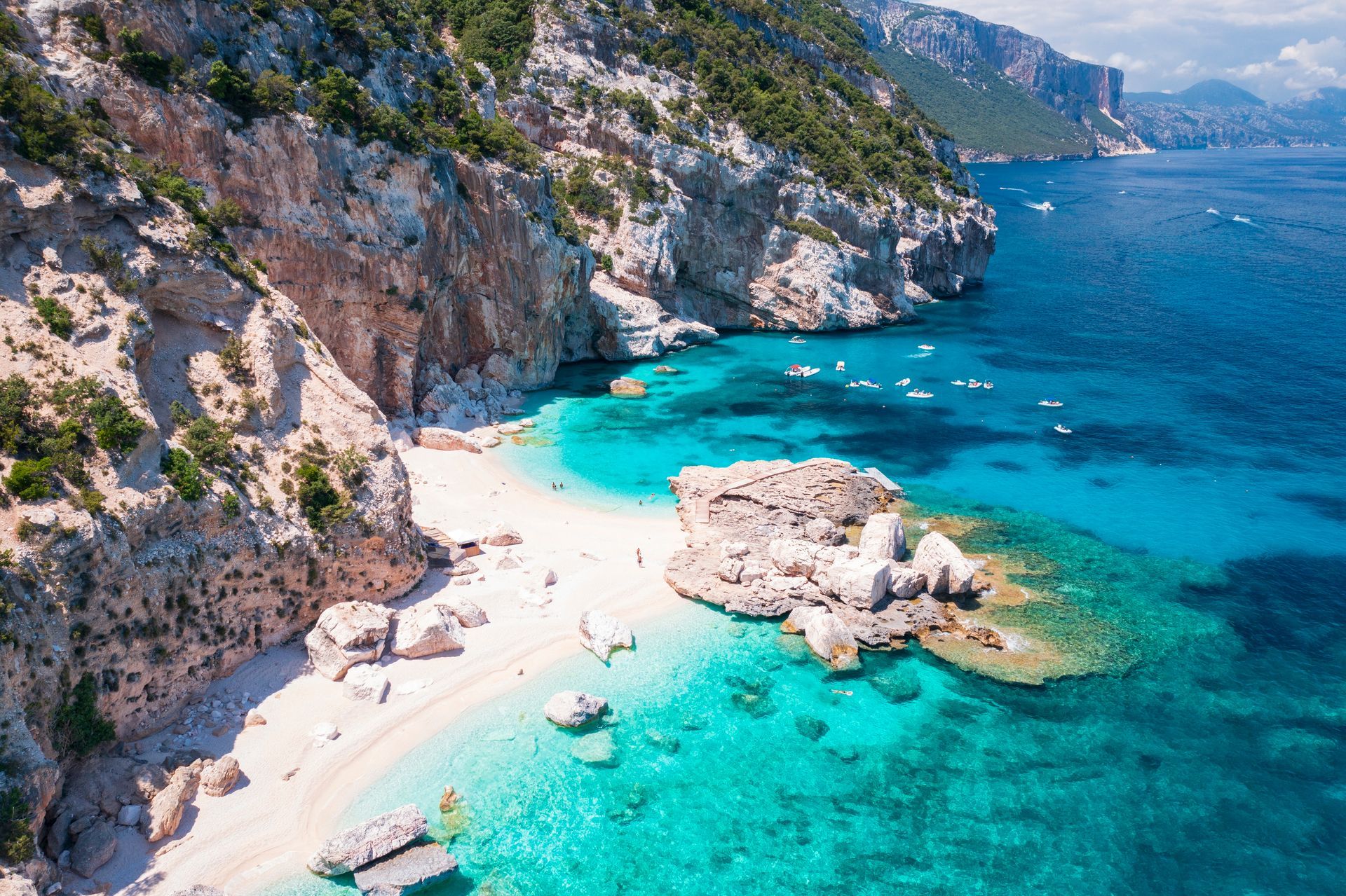 Sunny rocky cove with turquoise water, white sand beach, and small boats near steep cliffs
