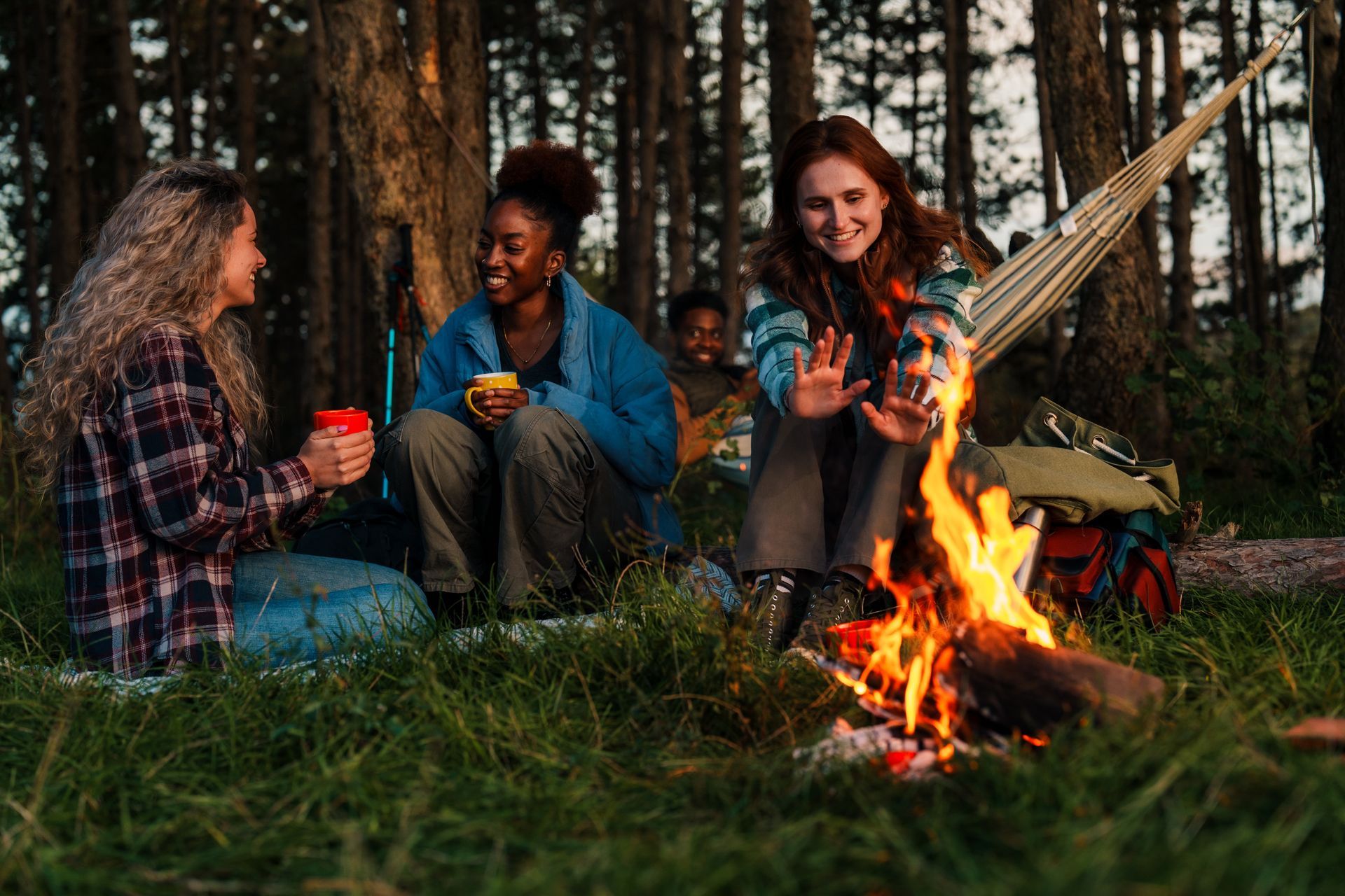 Friends sitting by a campfire in a forest, chatting and holding mugs