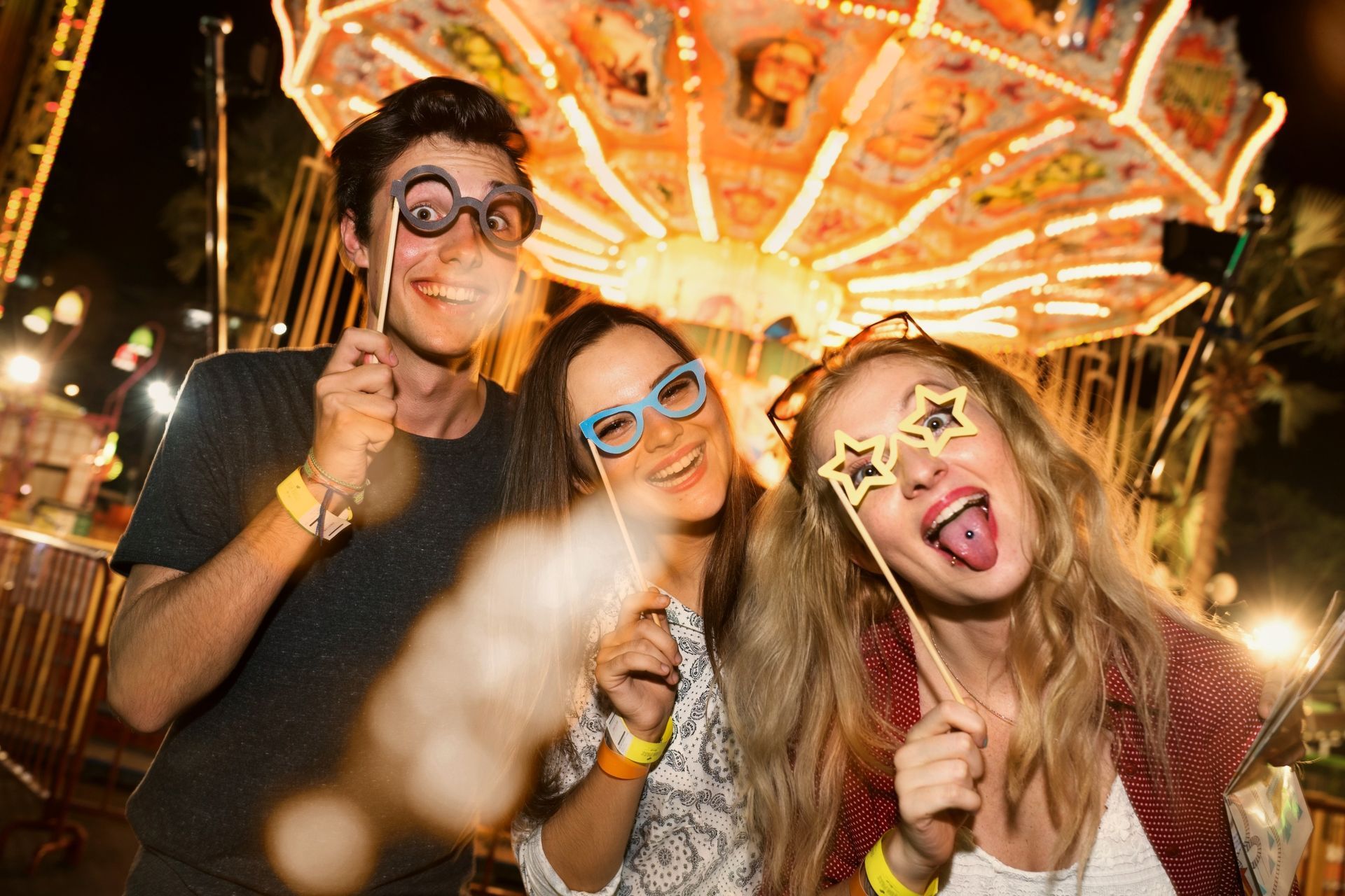 Three friends in party glasses posing at a brightly lit carnival ride at night